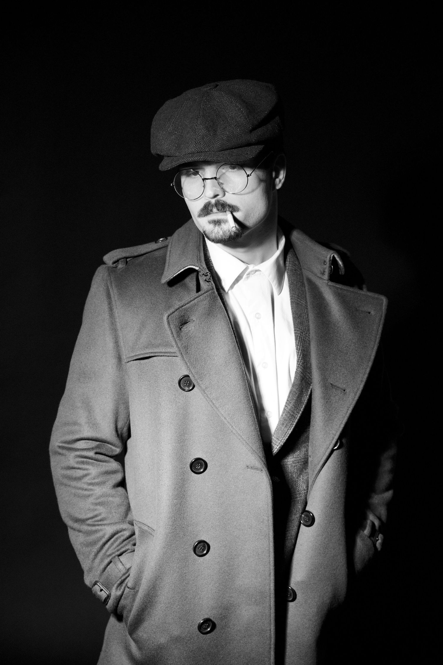Vintage-style black and white portrait of a man in trench coat, flat cap, and round glasses on dark background.