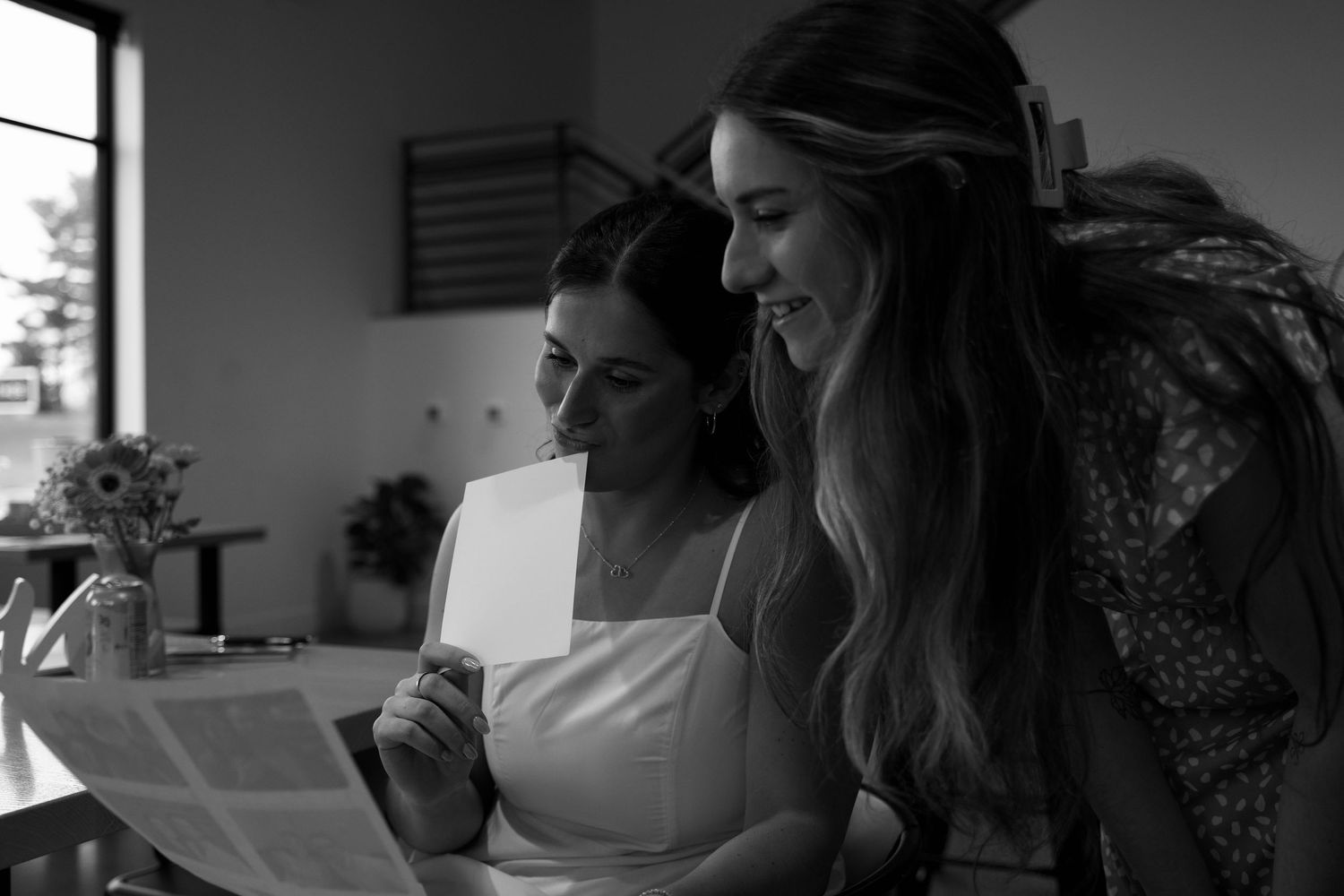 Black and white photo of two women smiling while reading a card together indoors.