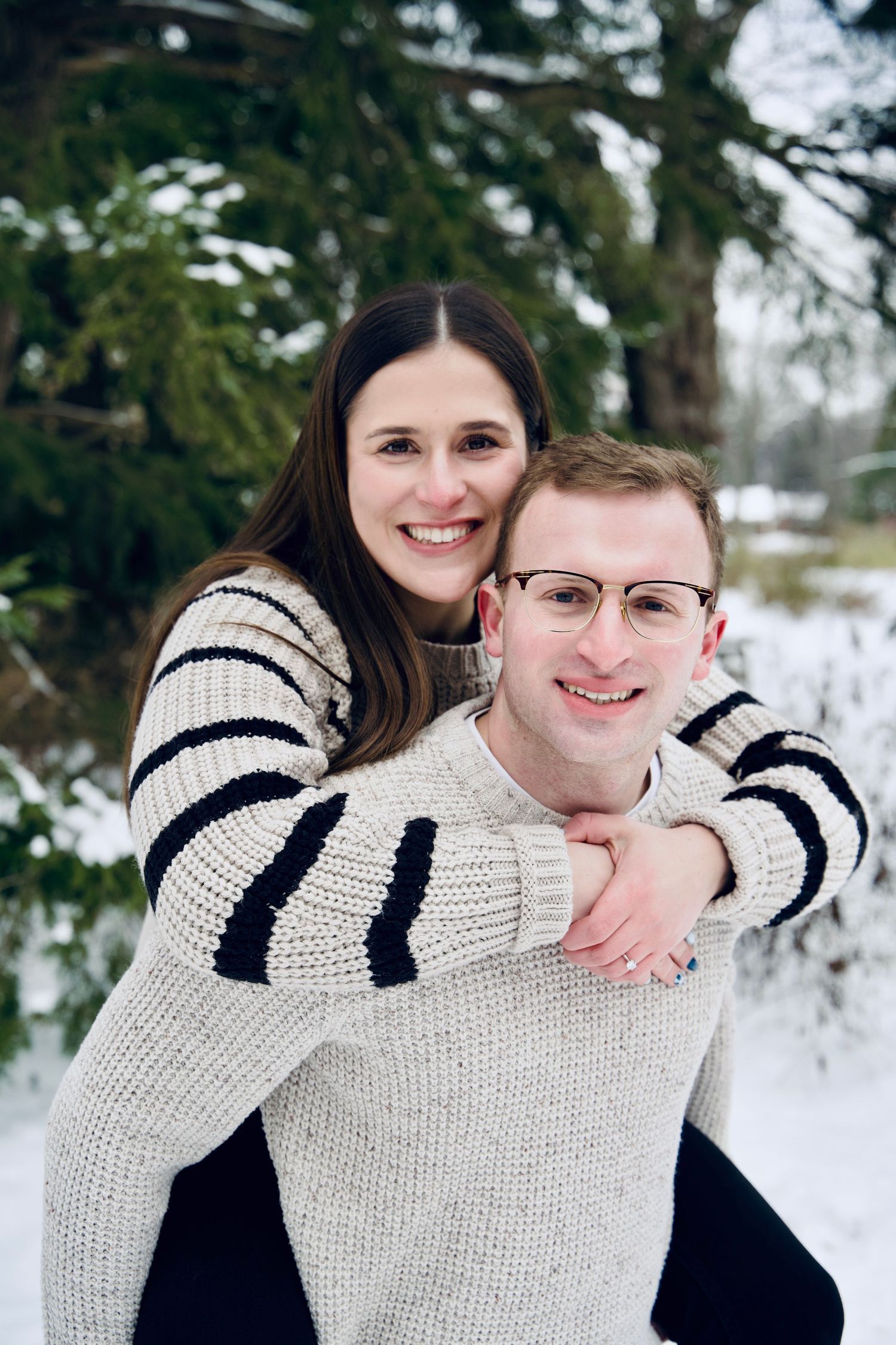 Happy couple in cozy knit sweaters share a piggyback pose surrounded by snow-covered evergreen trees.