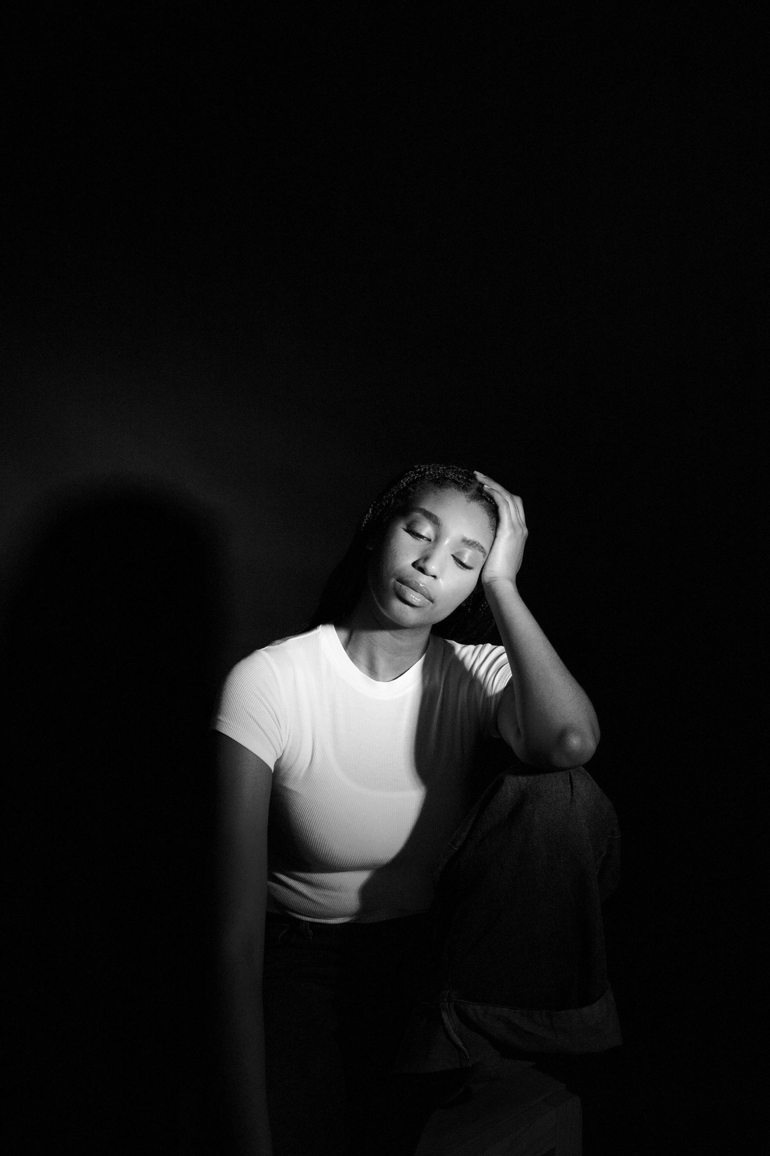 Black and white portrait of a young woman sitting against dark background with eyes closed, hand resting on her head.