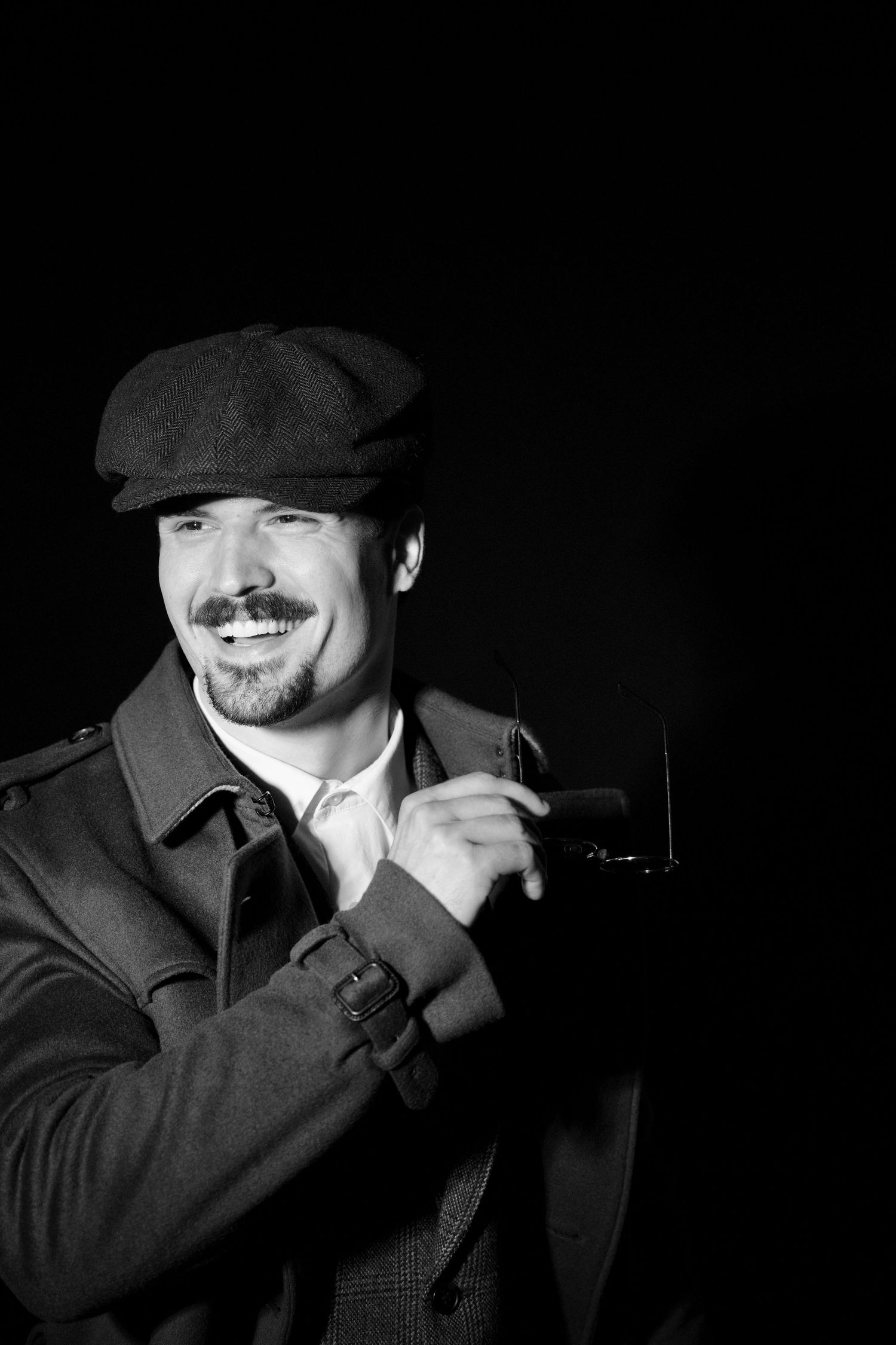 Smiling man in retro flat cap and trench coat holding glasses in a dramatic black and white studio portrait.