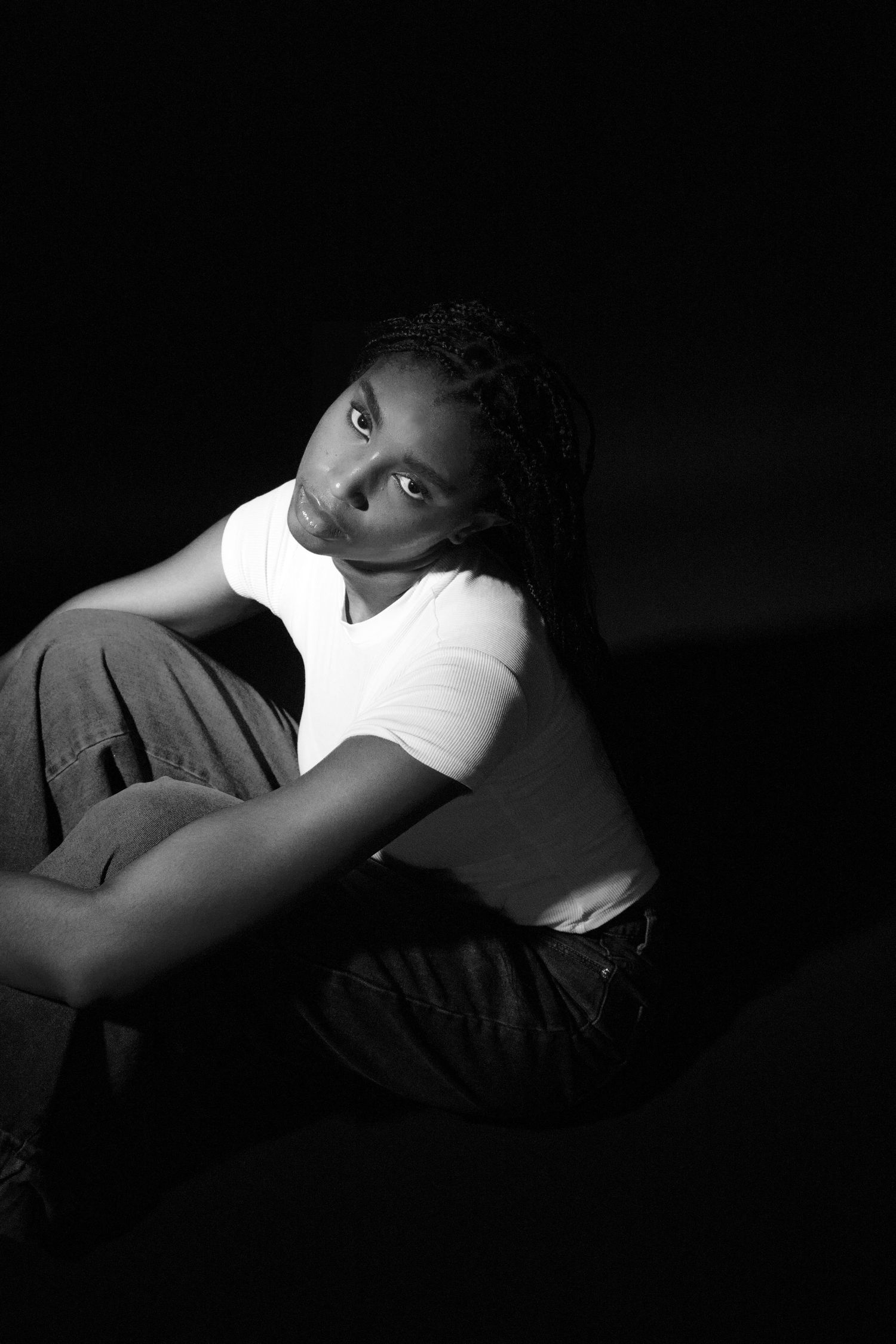 Black and white low-key portrait of a young woman in white t-shirt sitting casually on dark floor.