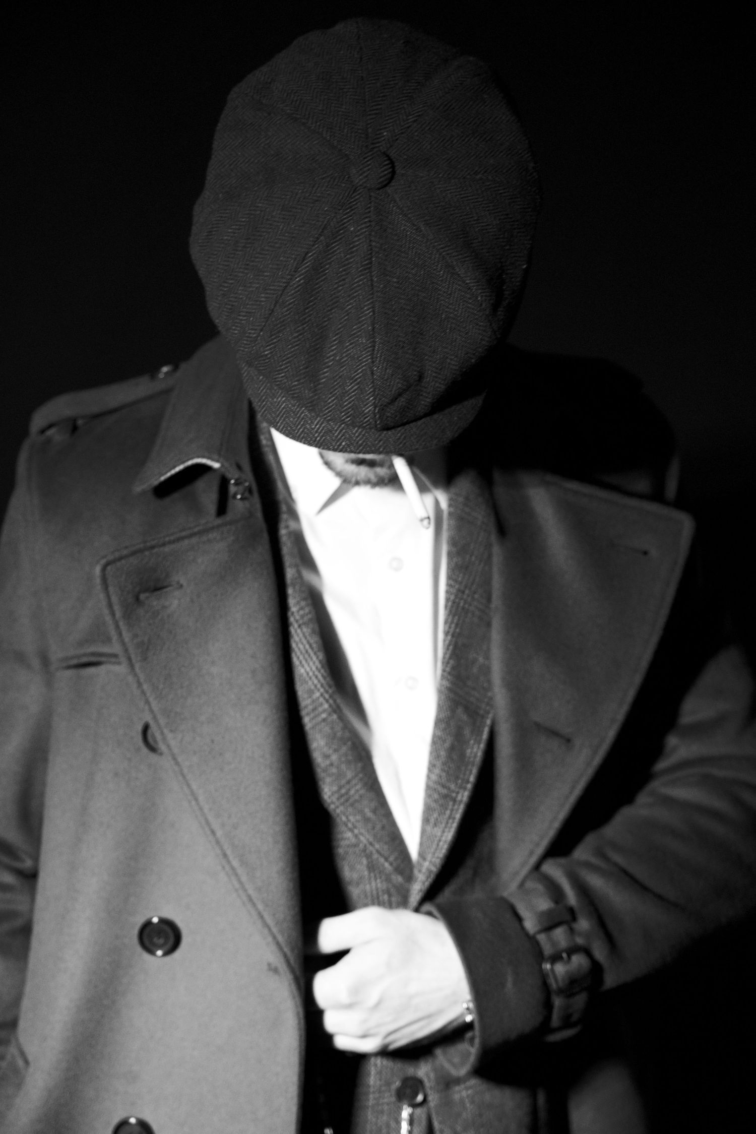 Mysterious black and white portrait of a man in trench coat and flat cap with face obscured in shadow.