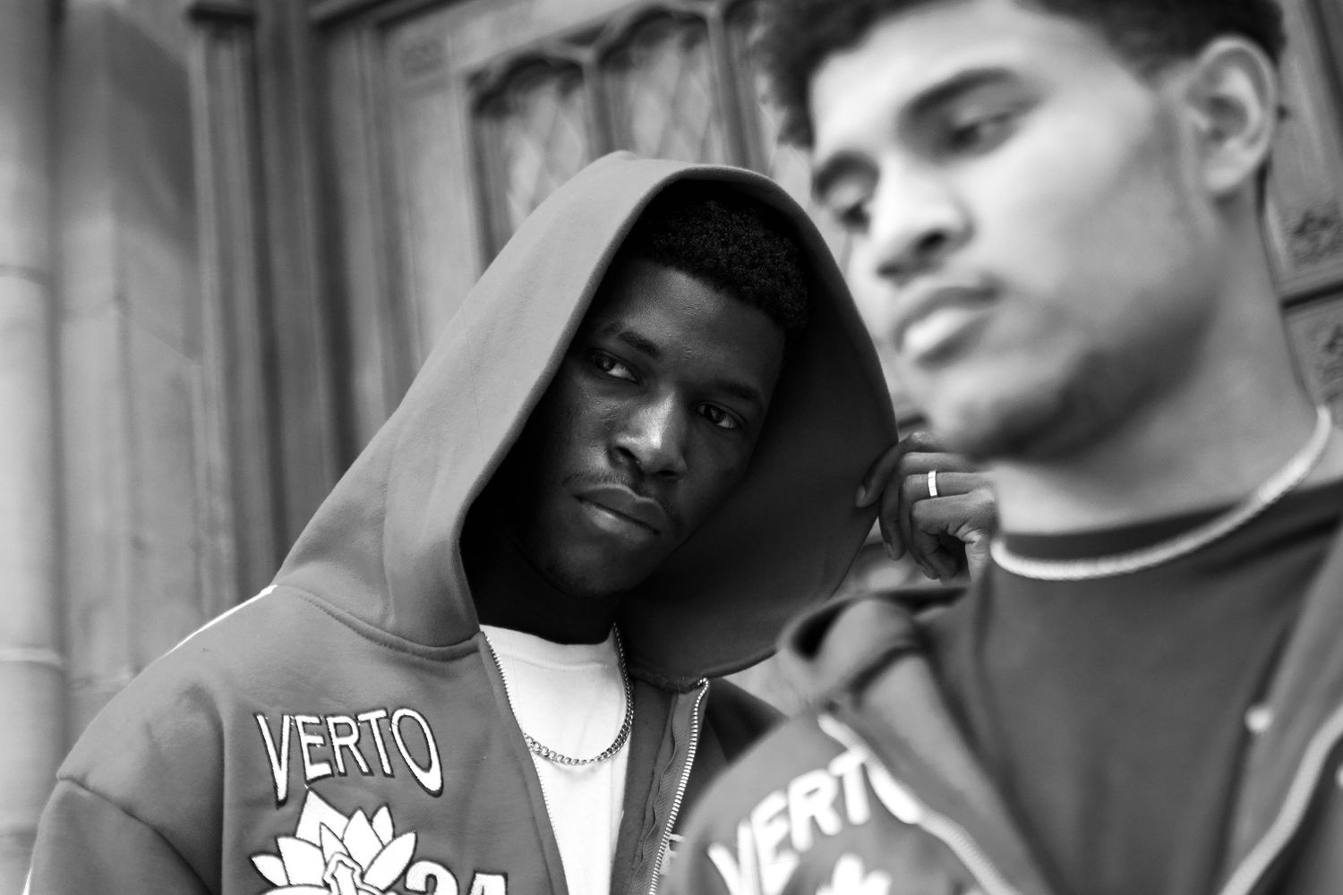 Black and white street portrait of two young men wearing matching Verto brand hoodies outdoors.