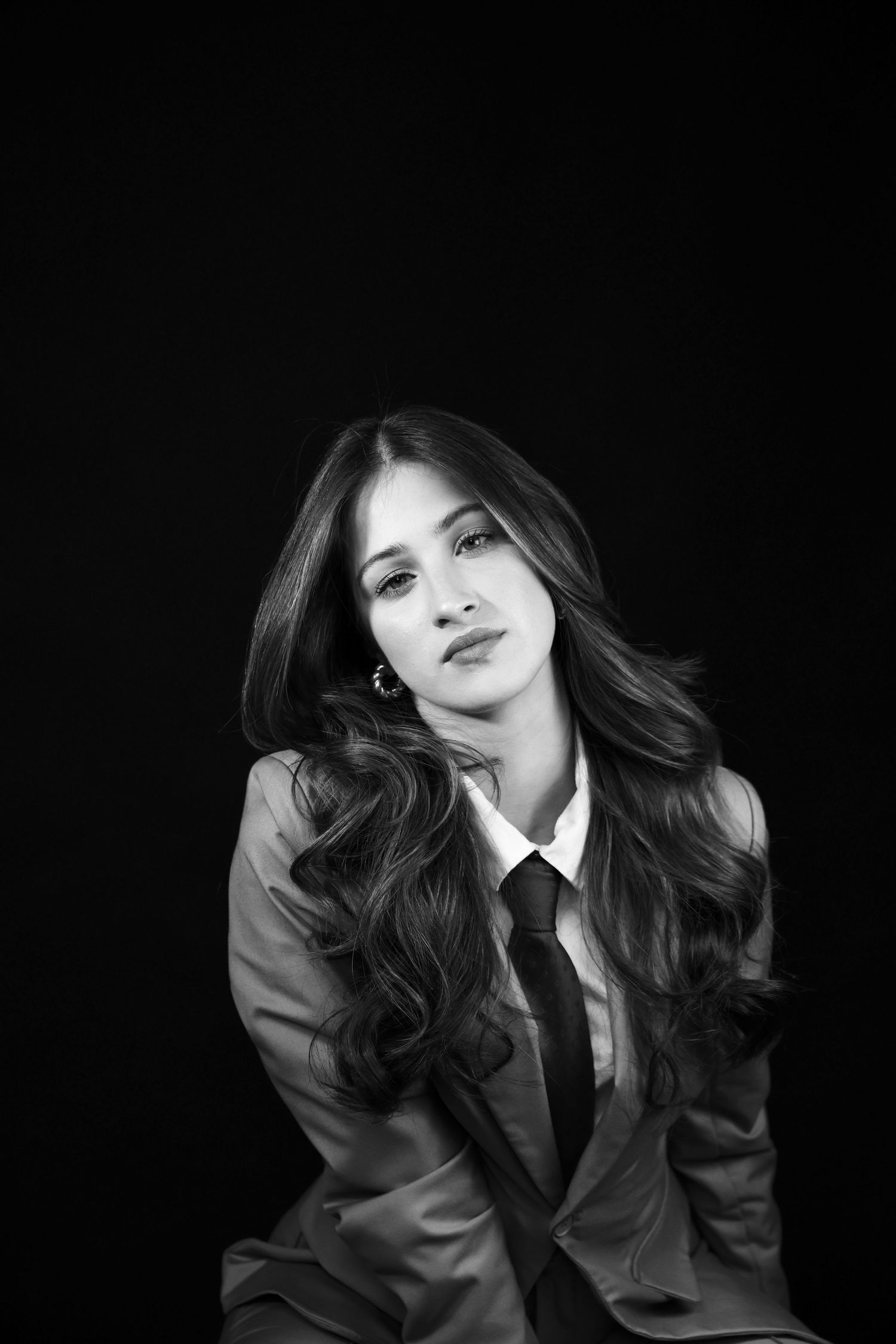 Black and white portrait of a young woman in a suit and tie with long wavy hair against dark background.