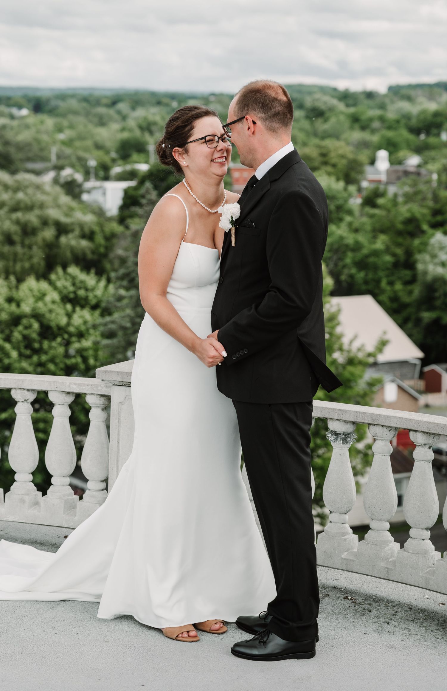 Bride in white gown and groom in black tuxedo share a romantic moment on a balcony with lush green trees behind.