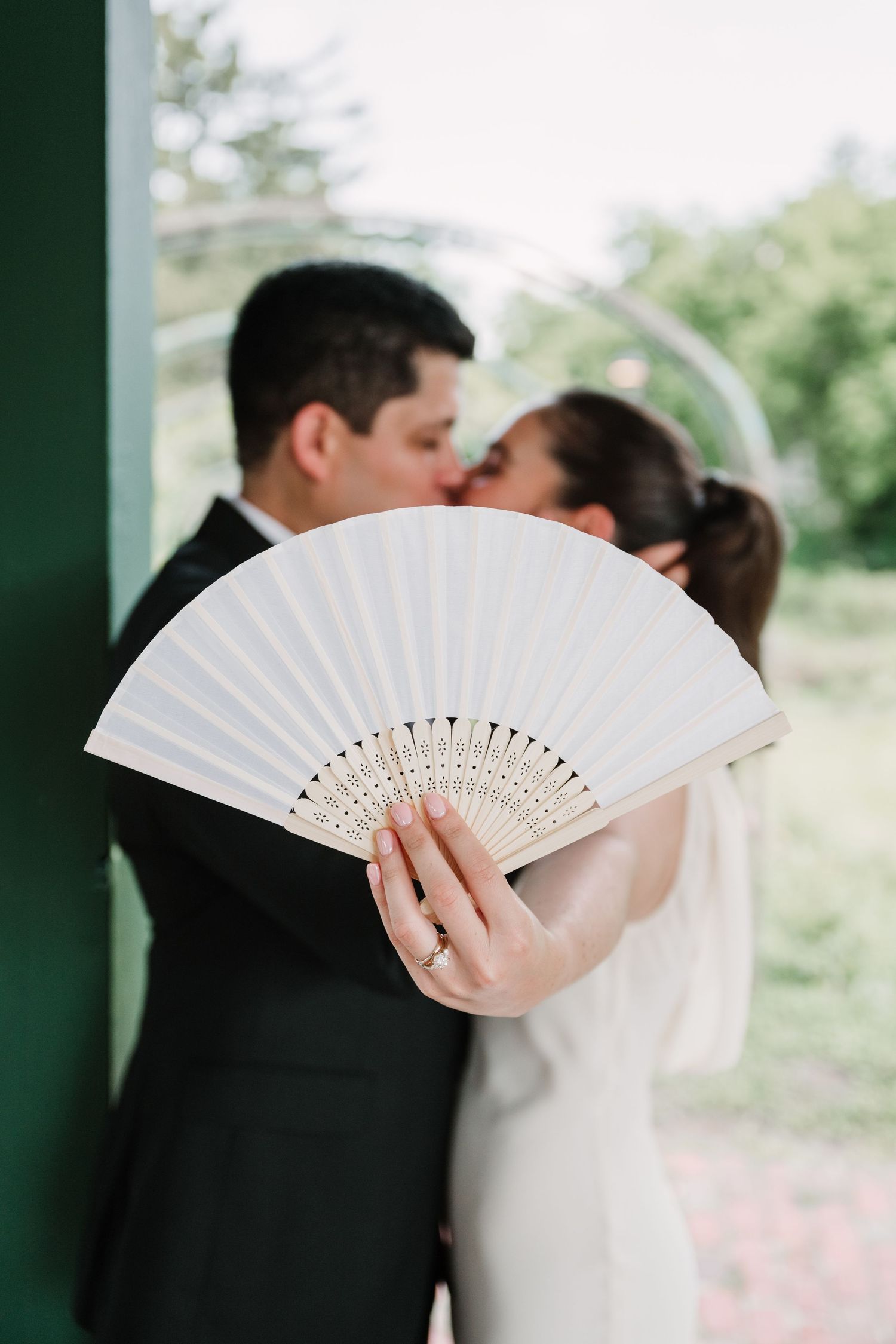 Romantic moment behind white decorative fan during outdoor wedding ceremony.