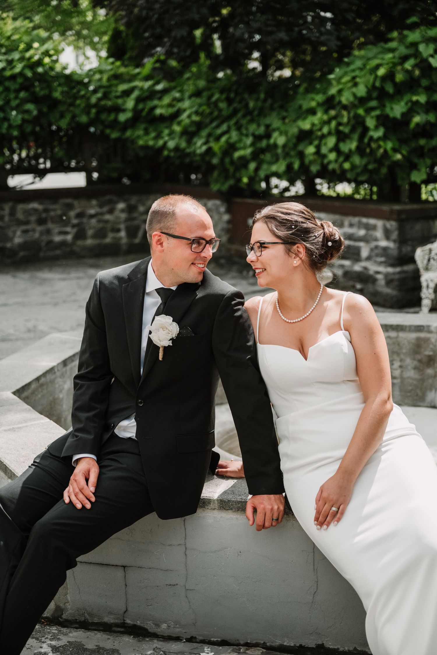 A bride and groom sit together on stone steps, holding hands and smiling at each other in an outdoor garden setting.