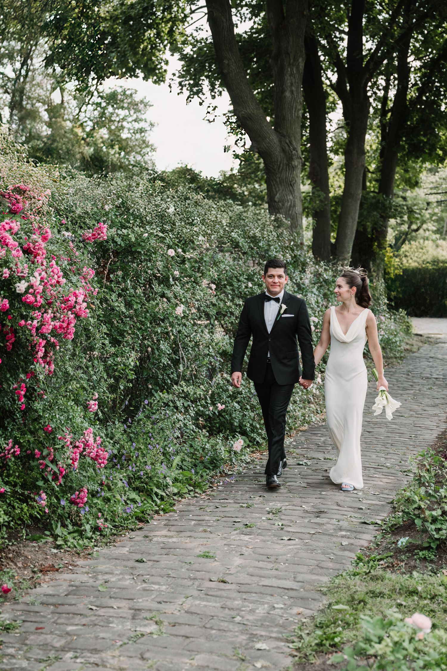 A couple walks along a cobblestone path lined with blooming pink roses.