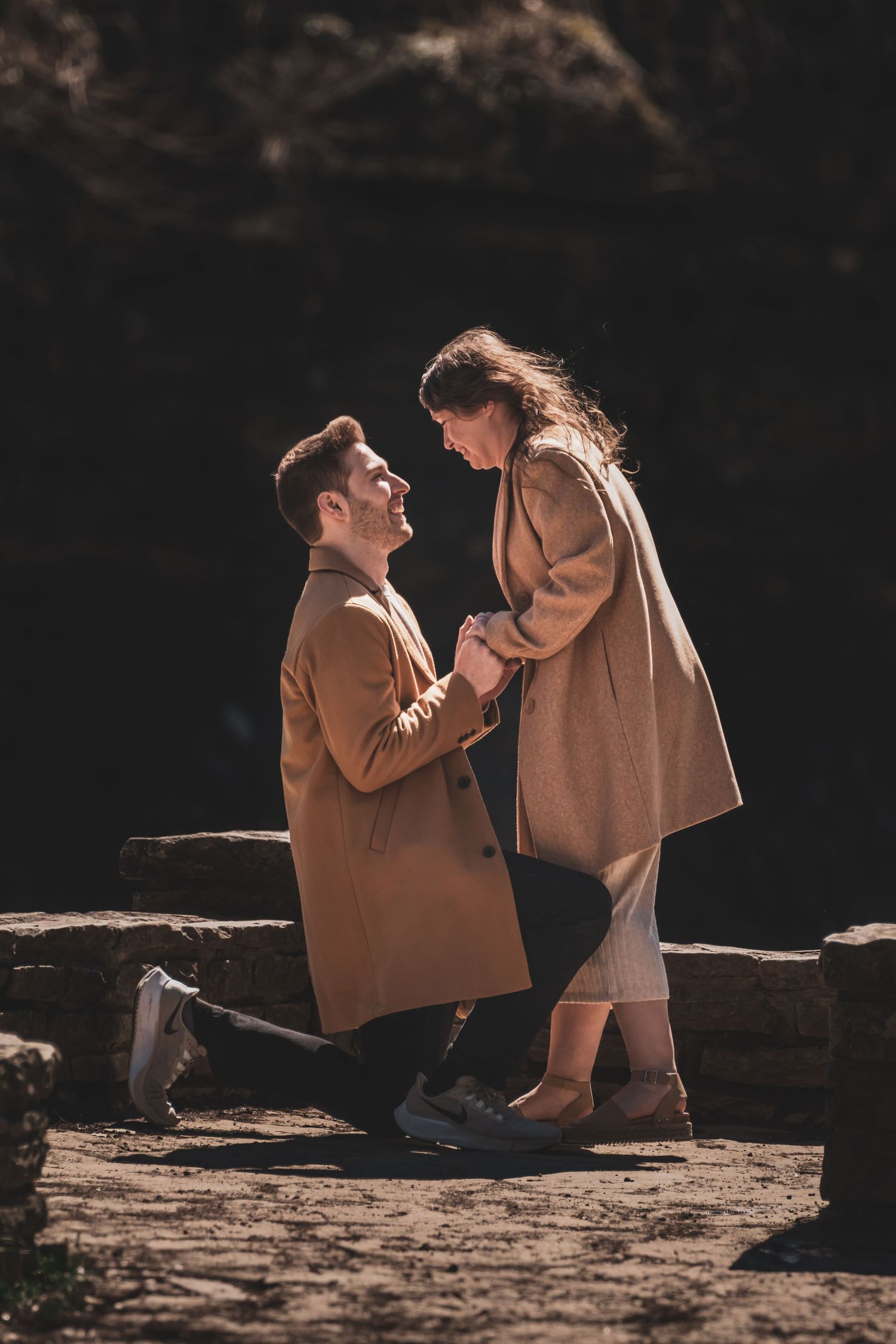 A couple in earth-toned clothing shares tender moments on rocky terrain in dramatic lighting.