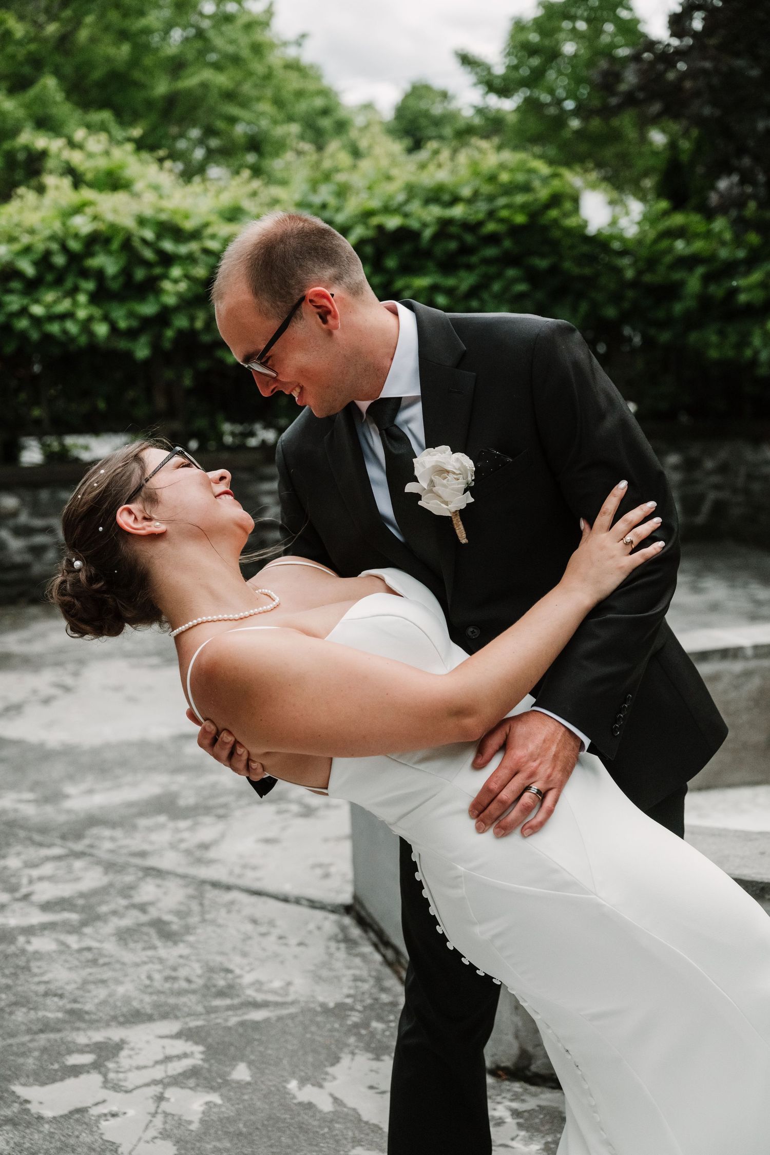 A groom dips his bride in a romantic pose outdoors, both wearing wedding attire with greenery in the background.