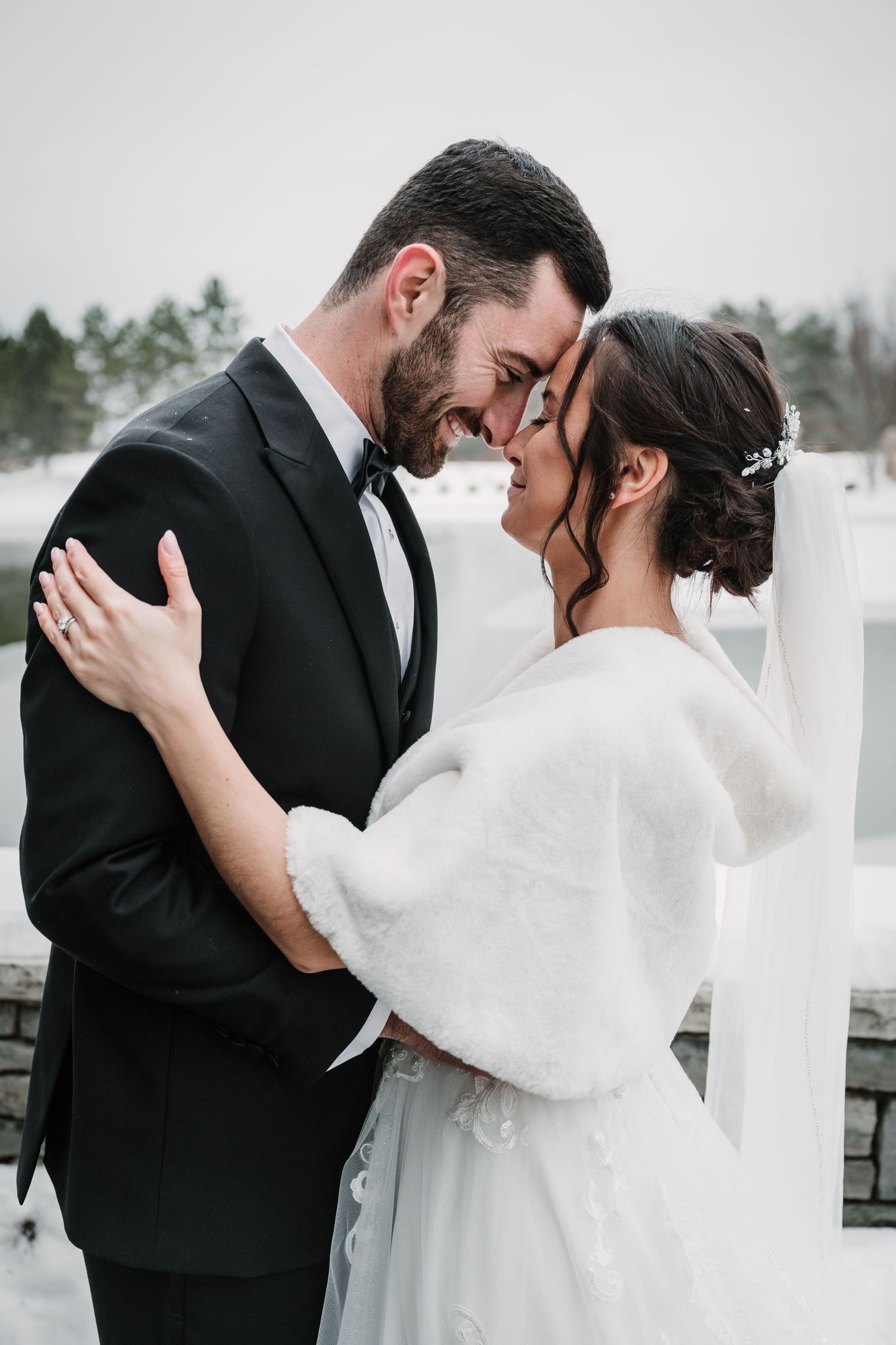 A winter wedding couple shares tender moments in a snowy setting with white fencing.