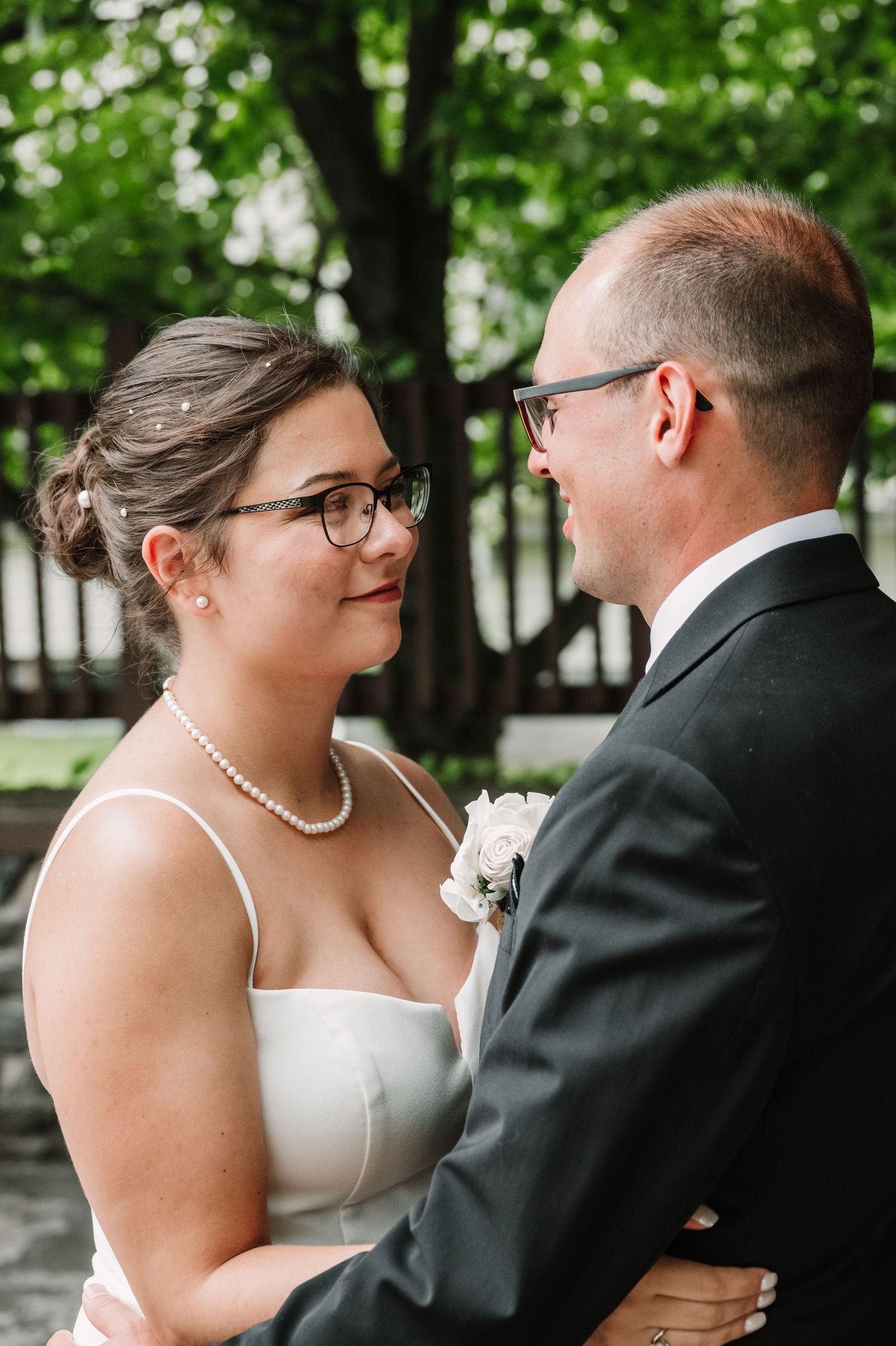 A bride and groom in wedding attire share a tender moment, both wearing glasses, outdoors with green trees in the background.