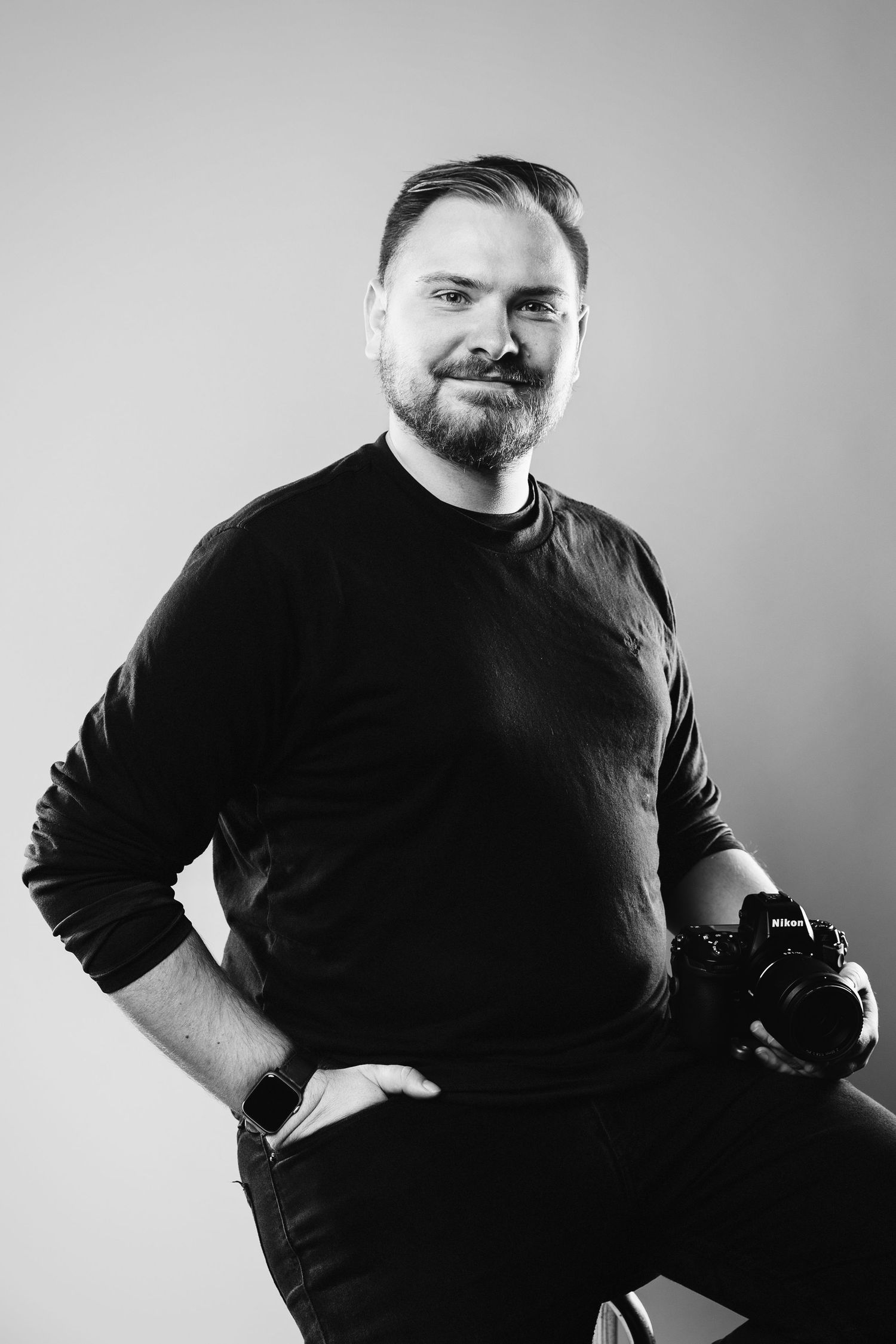 Professional black and white portrait of photographer holding camera against neutral background.