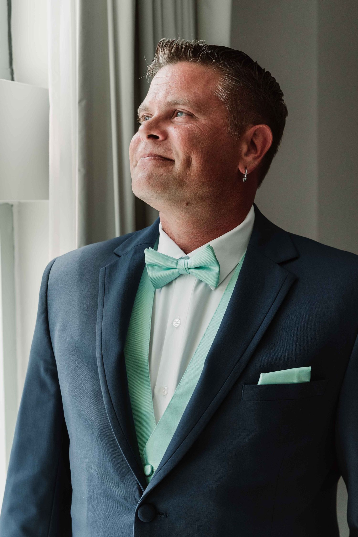 Groom in navy suit with mint bowtie poses by window in natural light.