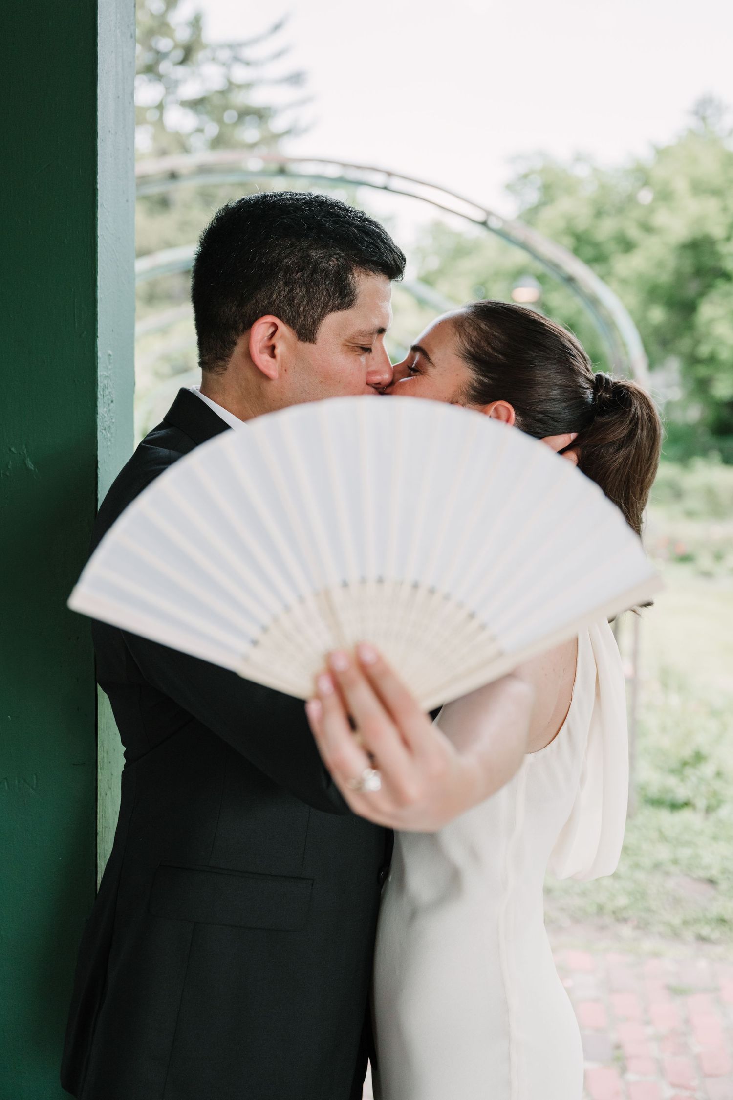 A romantic wedding couple shares a kiss behind a white folding fan in an outdoor garden setting.