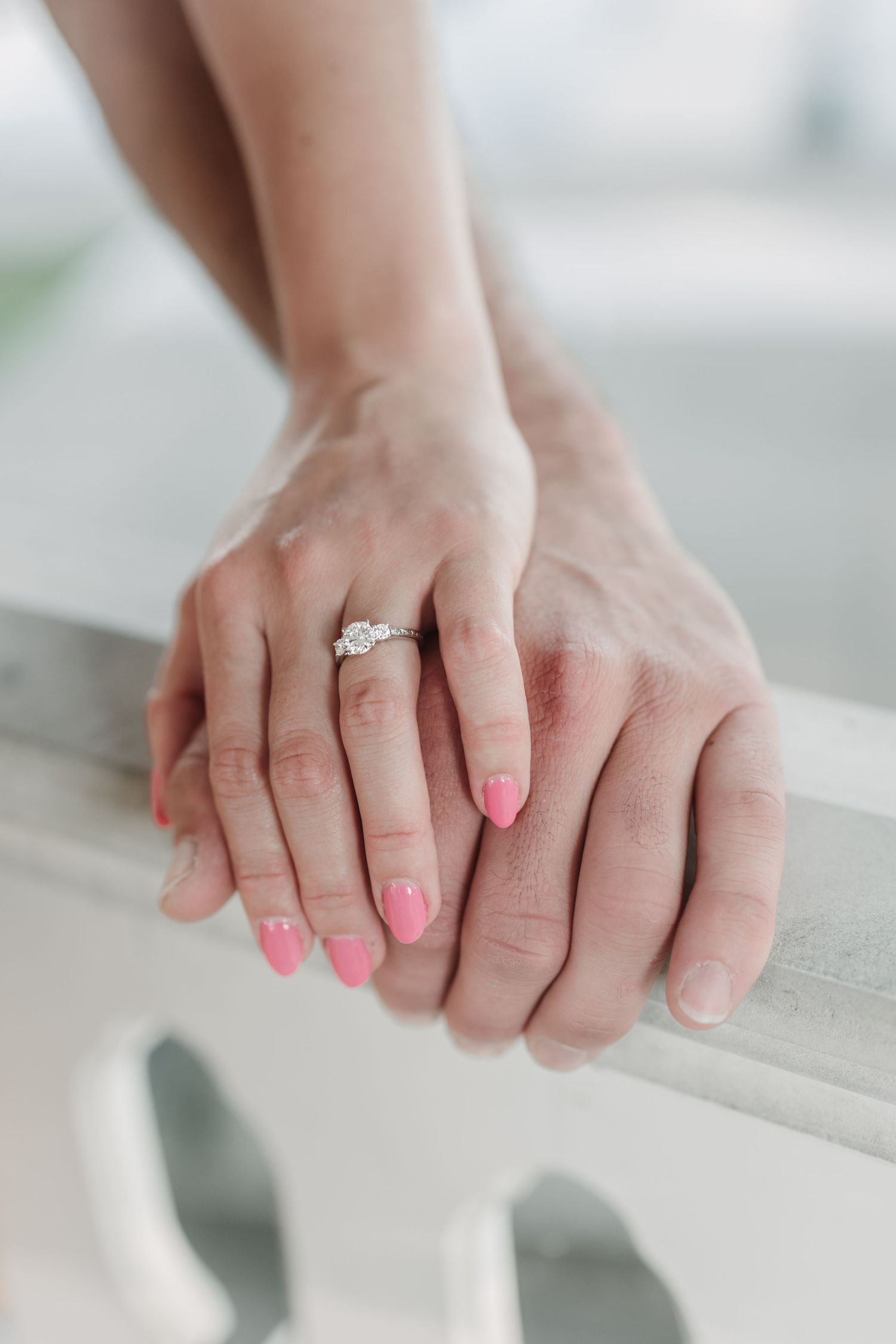 Two hands intertwined on a white railing, showcasing a sparkling engagement ring.