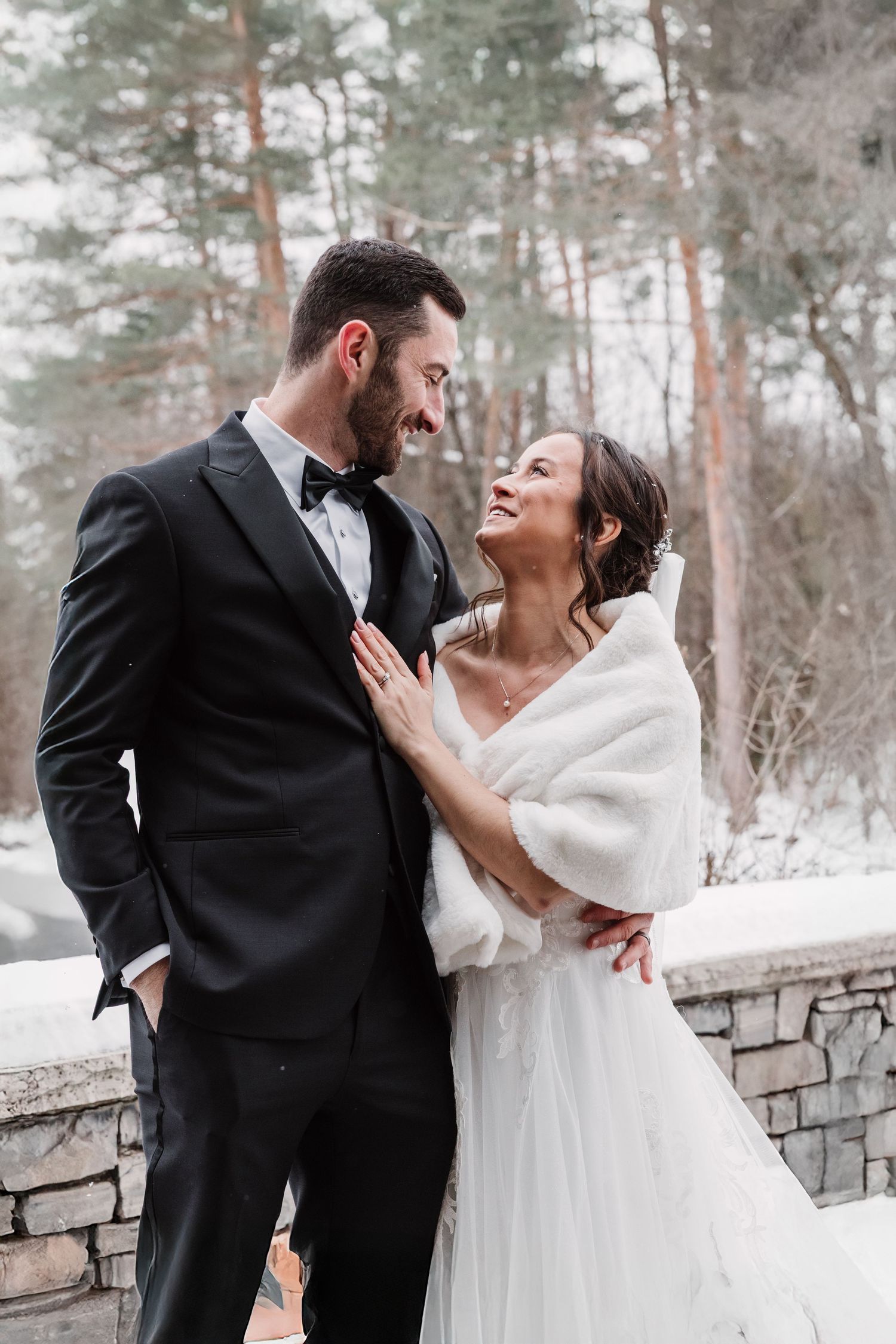 A winter wedding couple shares joyful moments against a snowy stone wall backdrop.