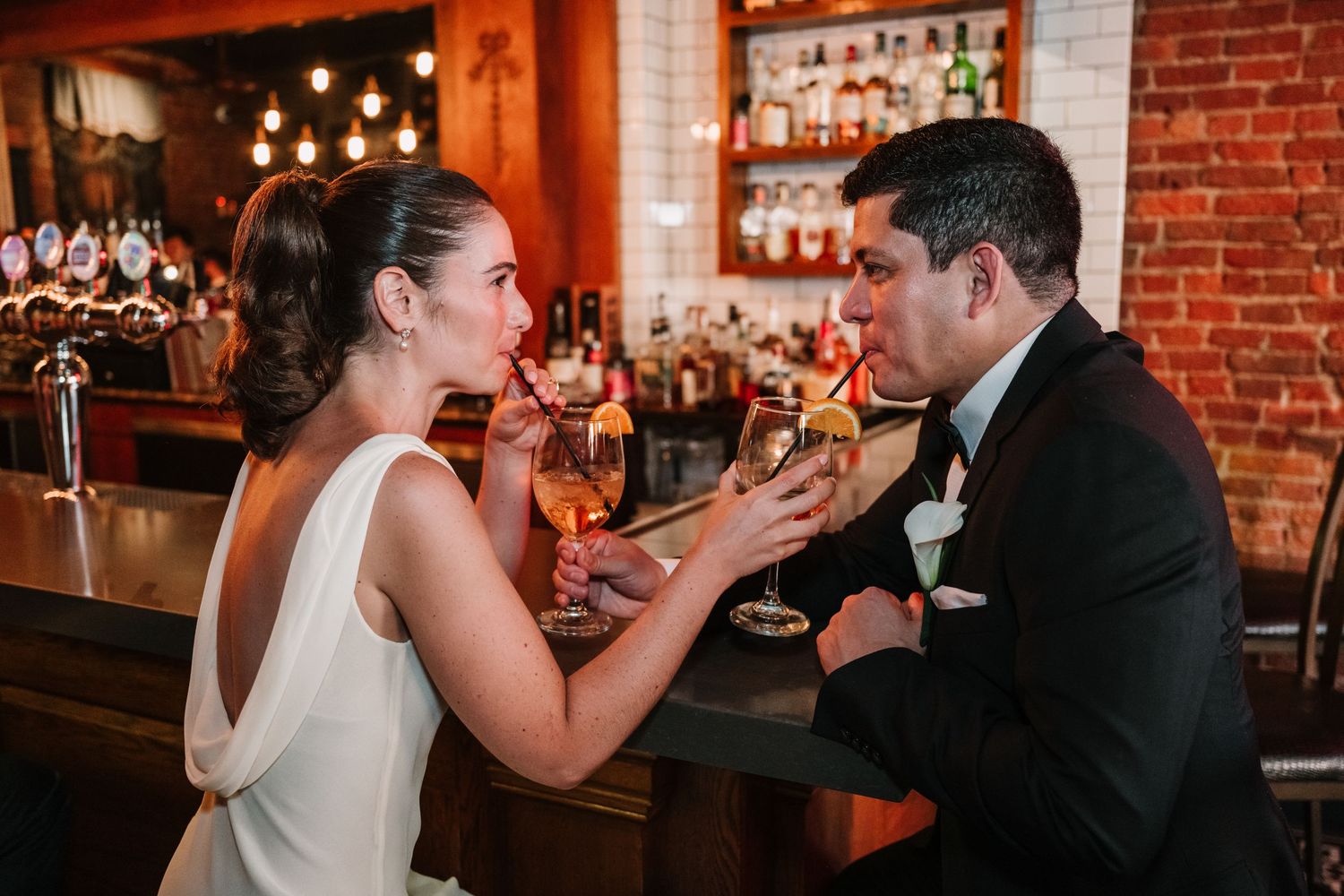 A couple shares a playful moment at a cozy bar with brick walls and ambient lighting.
