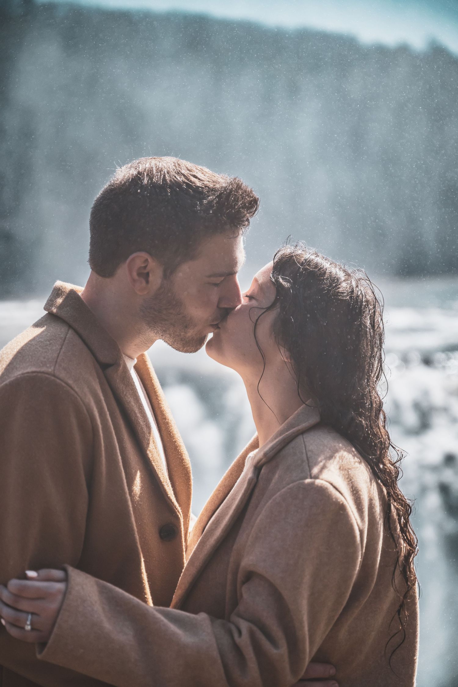 Romantic winter photoshoot featuring couple in tan coats against misty background.