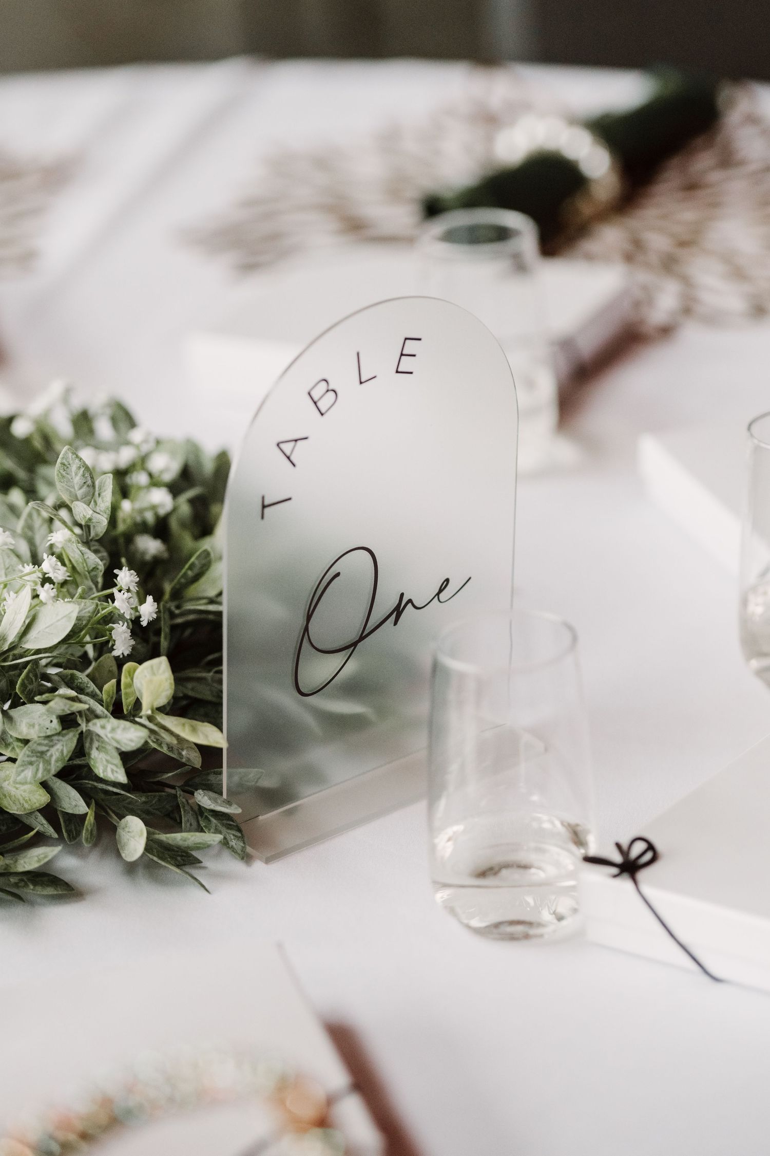 Elegant wedding table one sign on frosted acrylic arch display with greenery floral arrangement and glass bud vase.