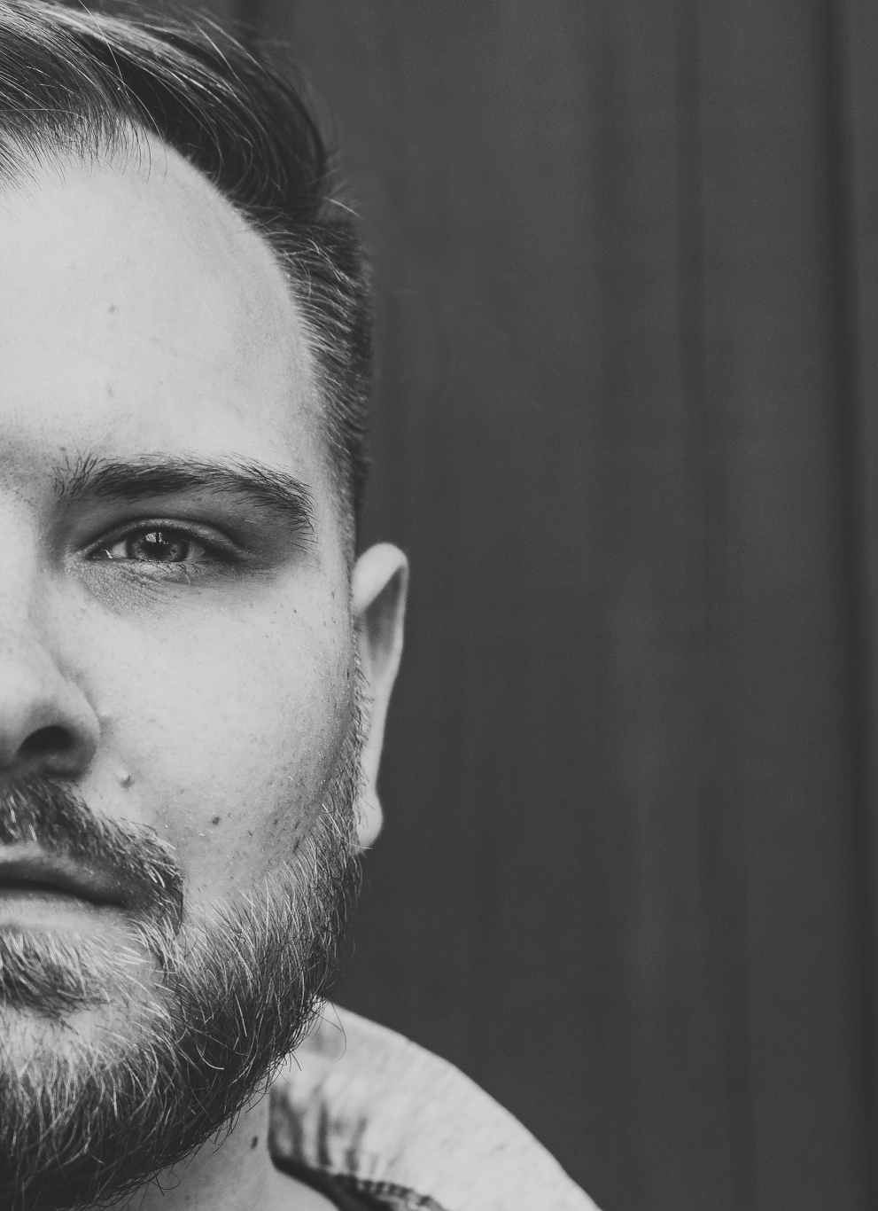 Dramatic black and white close-up portrait showing half of face with beard against dark background.