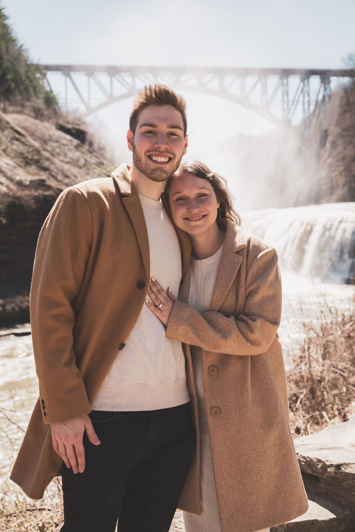 Couple in matching tan coats sharing intimate moments by a waterfall on a sunny winter day.