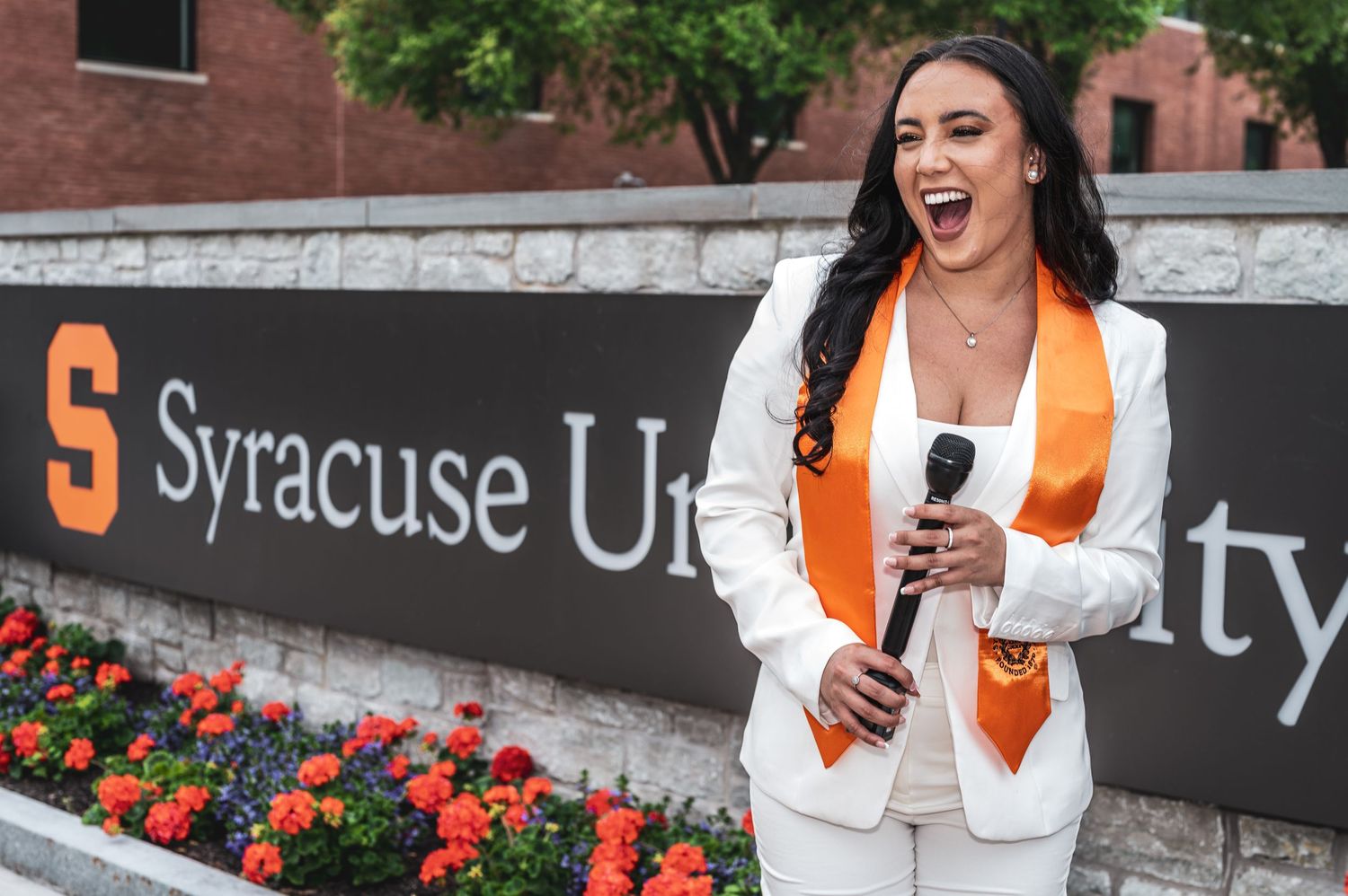 Graduate in white blazer and orange stole celebrating near Syracuse University sign.
