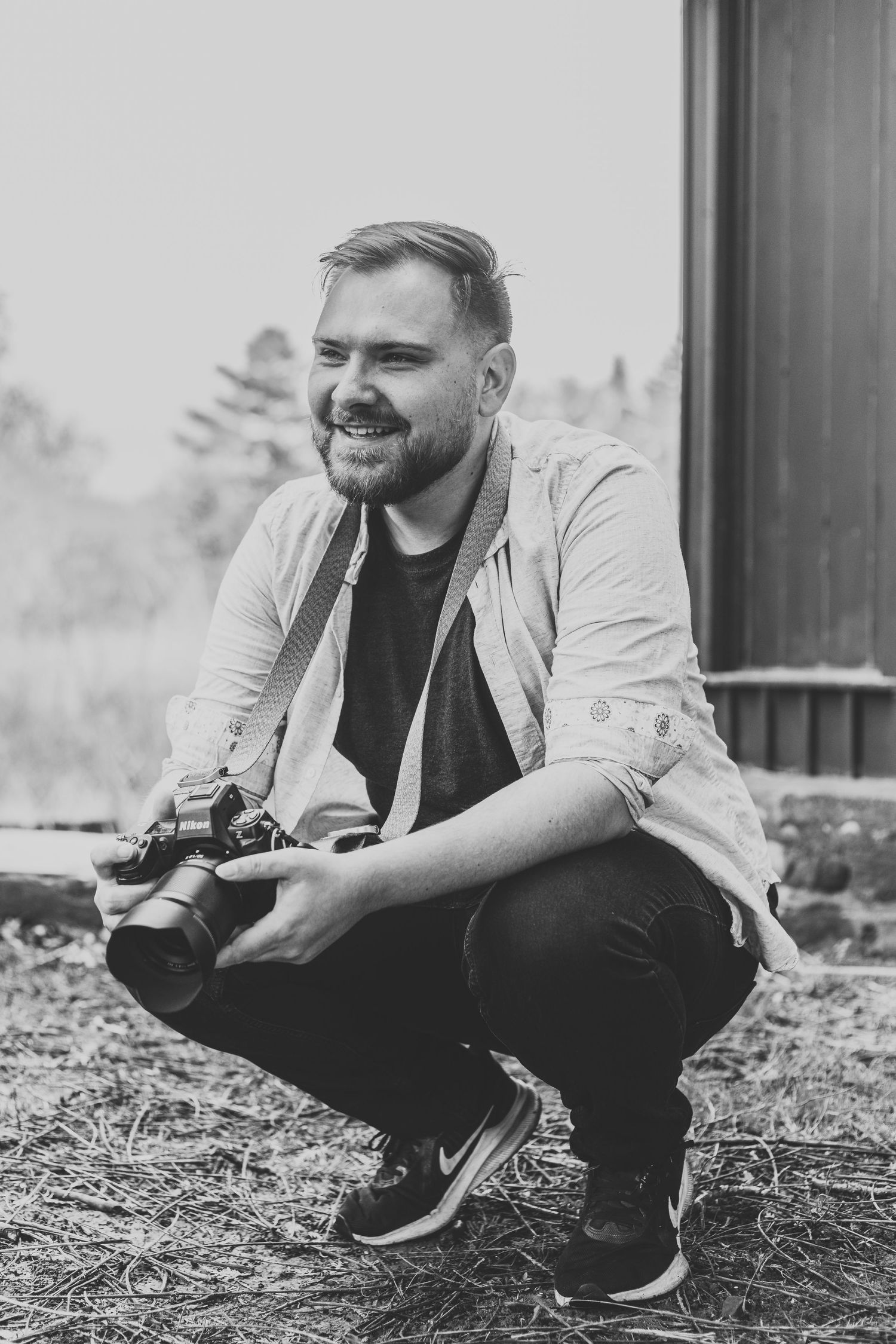 Black and white portrait of a photographer crouching outdoors with a camera.