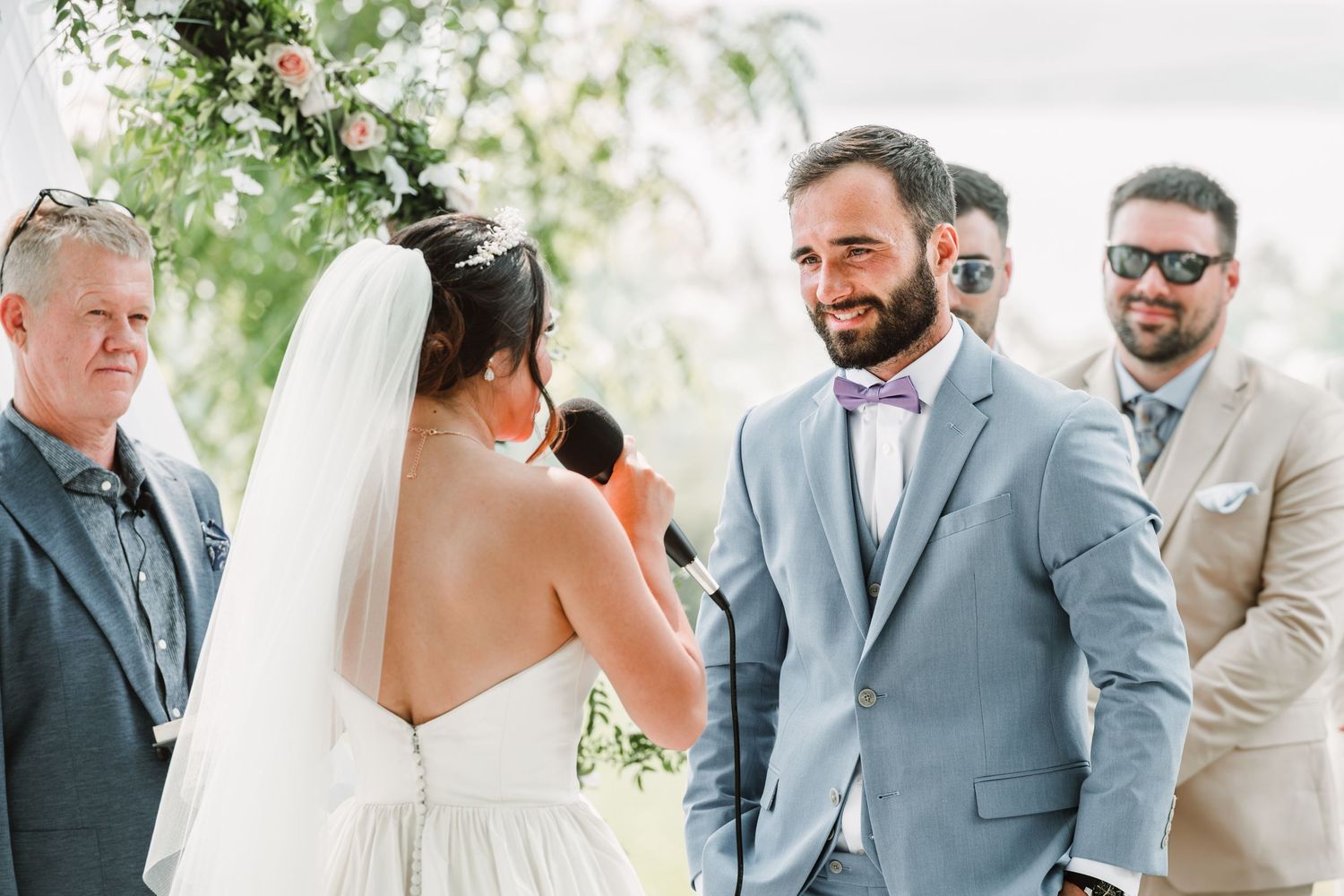 Wedding ceremony group shot with groomsmen in gray suits and white floral decorations.