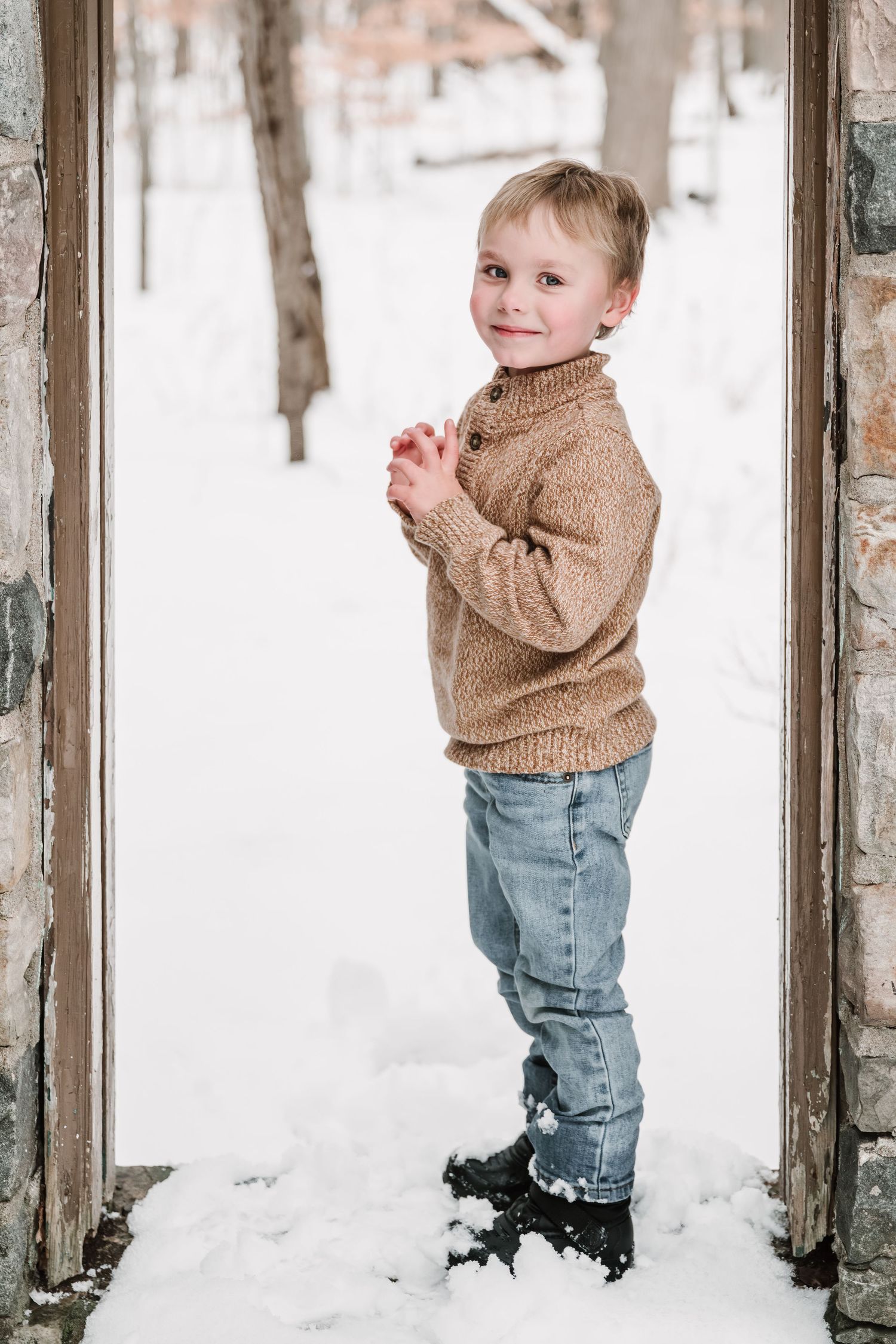 Toddler in tan sweater playing in snowy winter setting between rustic pillars.