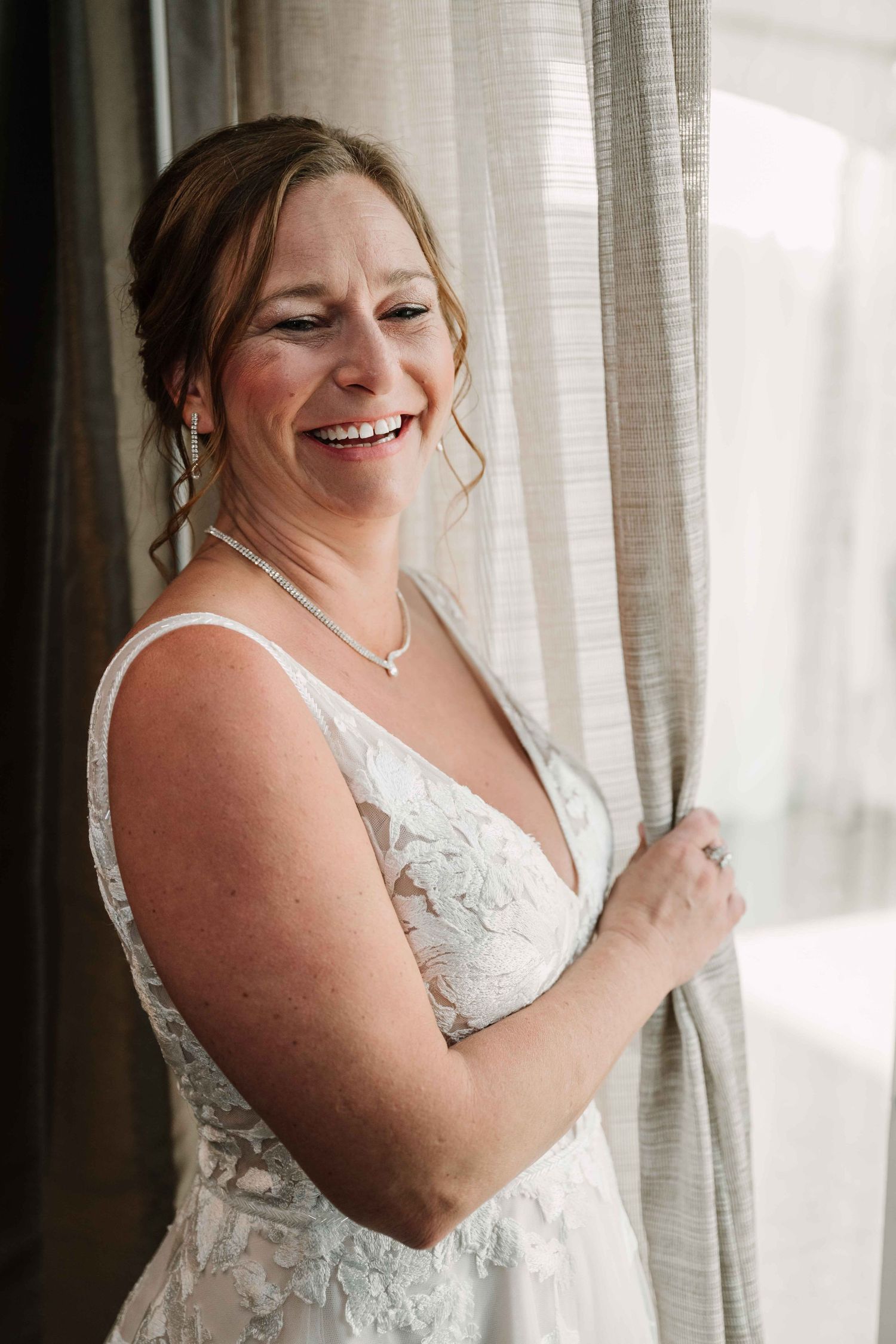 Candid moment of bride in lace dress laughing by window.