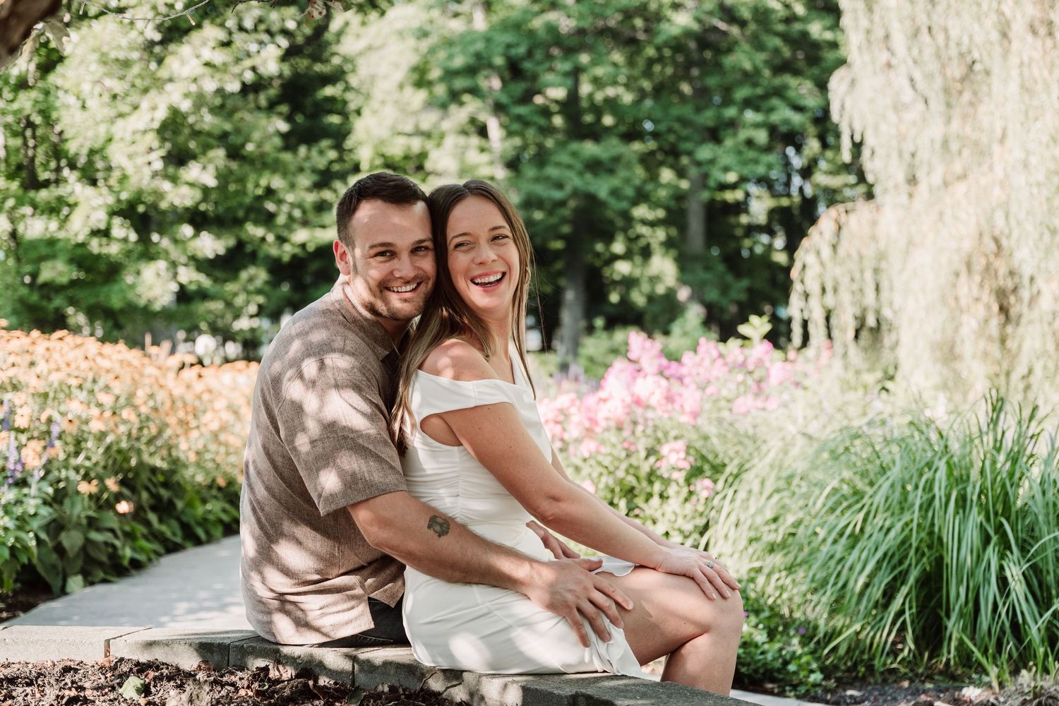 A happy couple sits together on a stone bench in a garden with blooming pink flowers and ornamental grasses.