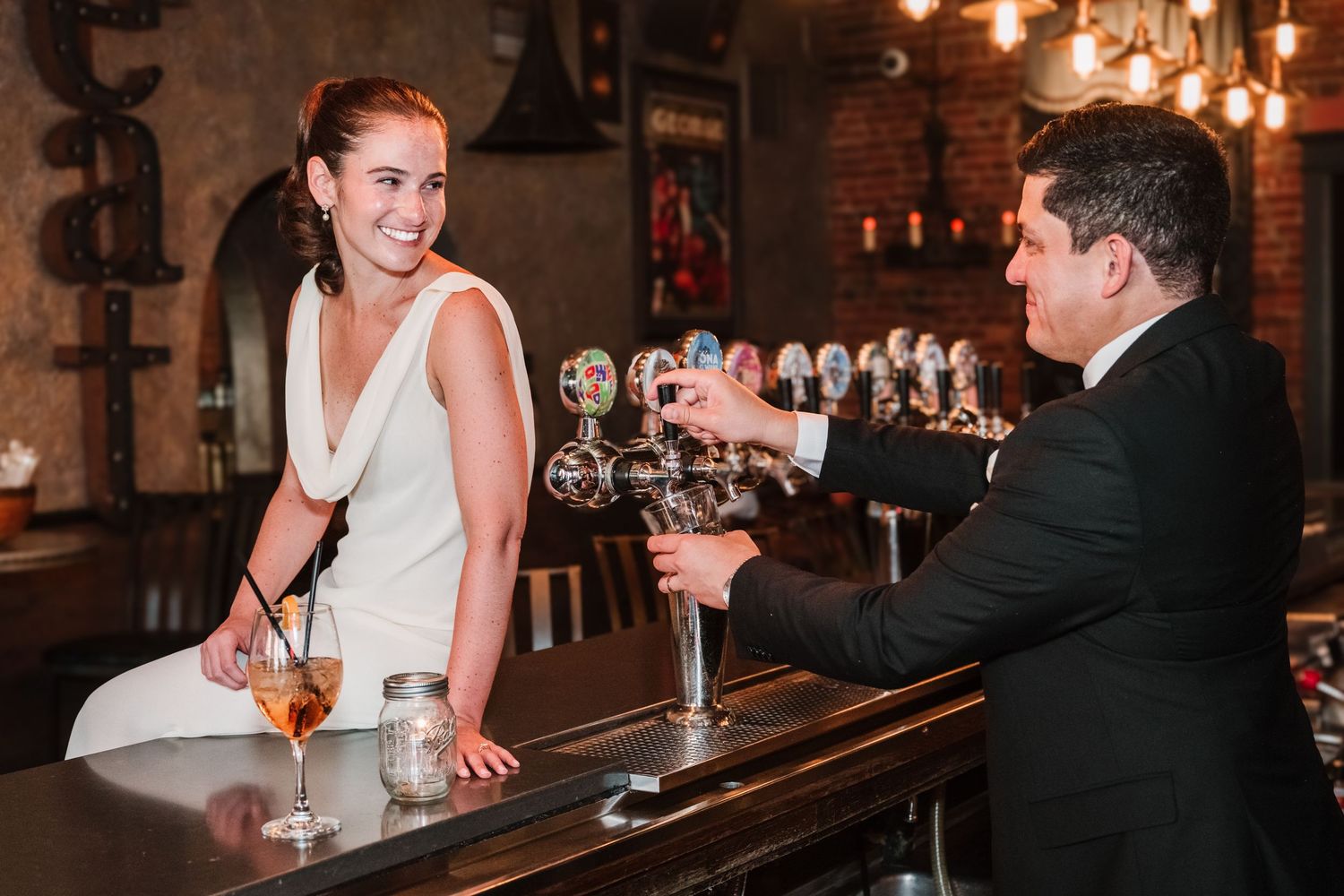 Wedding couple shares drinks and laughter at a rustic bar with ambient lighting.