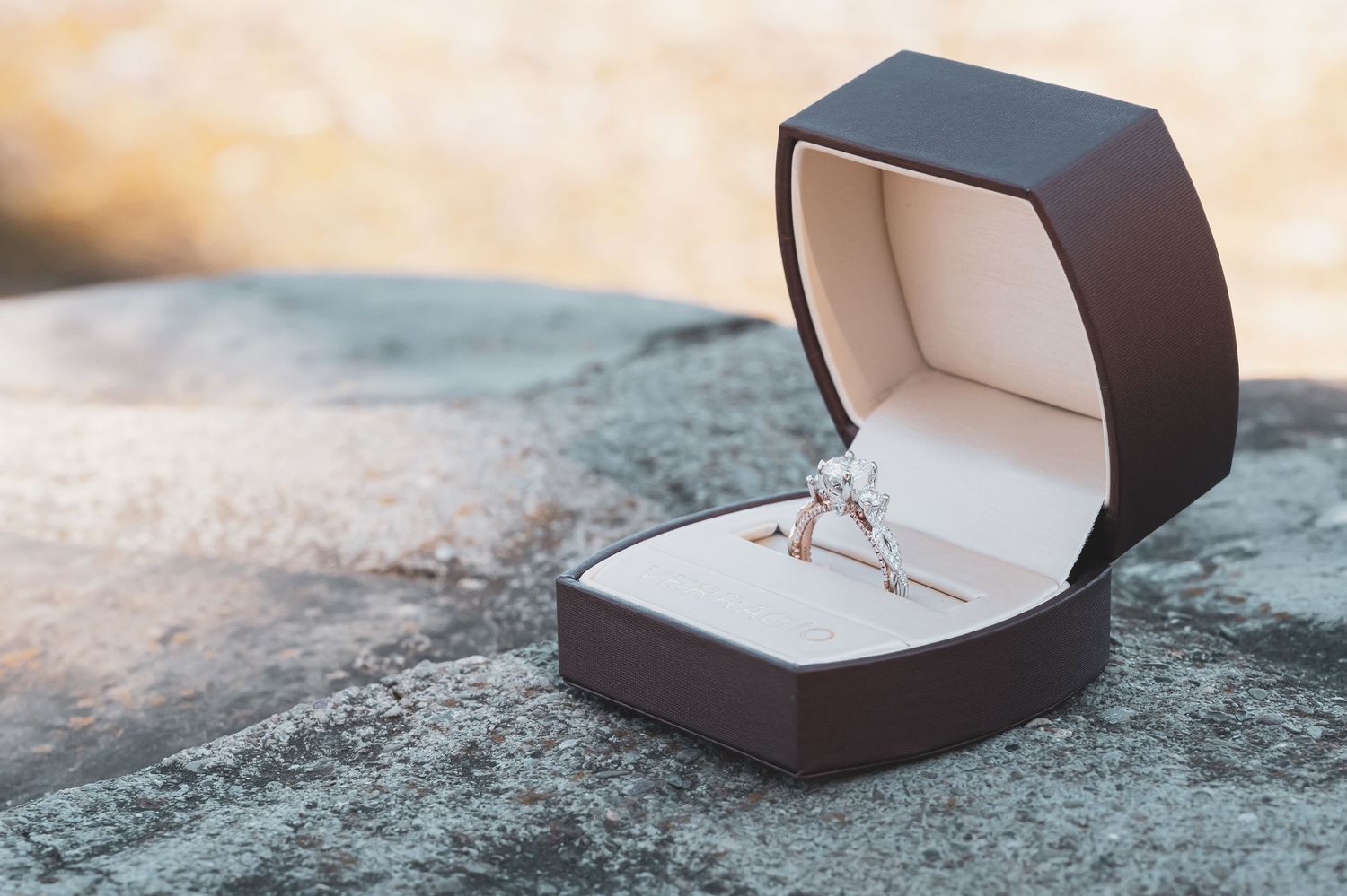 Black engagement ring box opens to reveal a sparkling diamond ring on a stone surface.