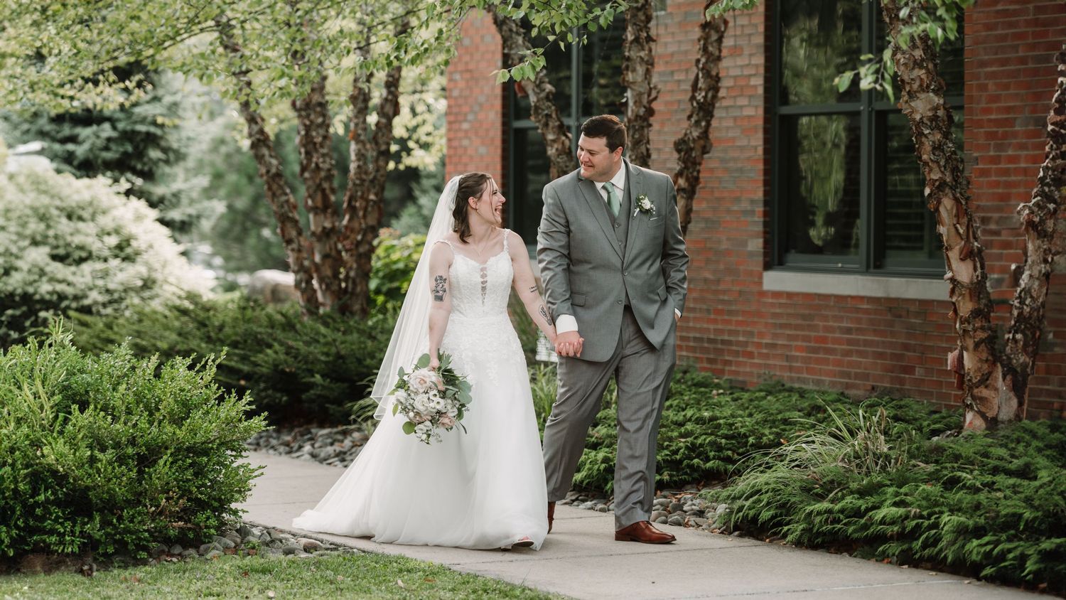 Wedding couple walking along a brick-lined pathway surrounded by lush landscaping.