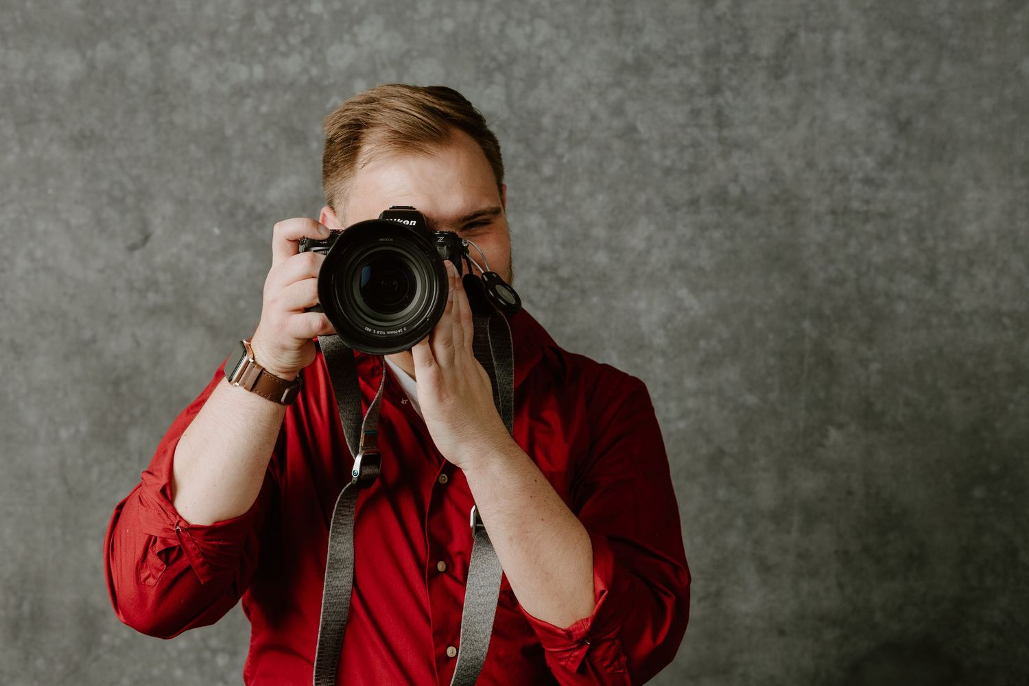 Photographer in red shirt capturing photo with professional DSLR camera against textured wall.