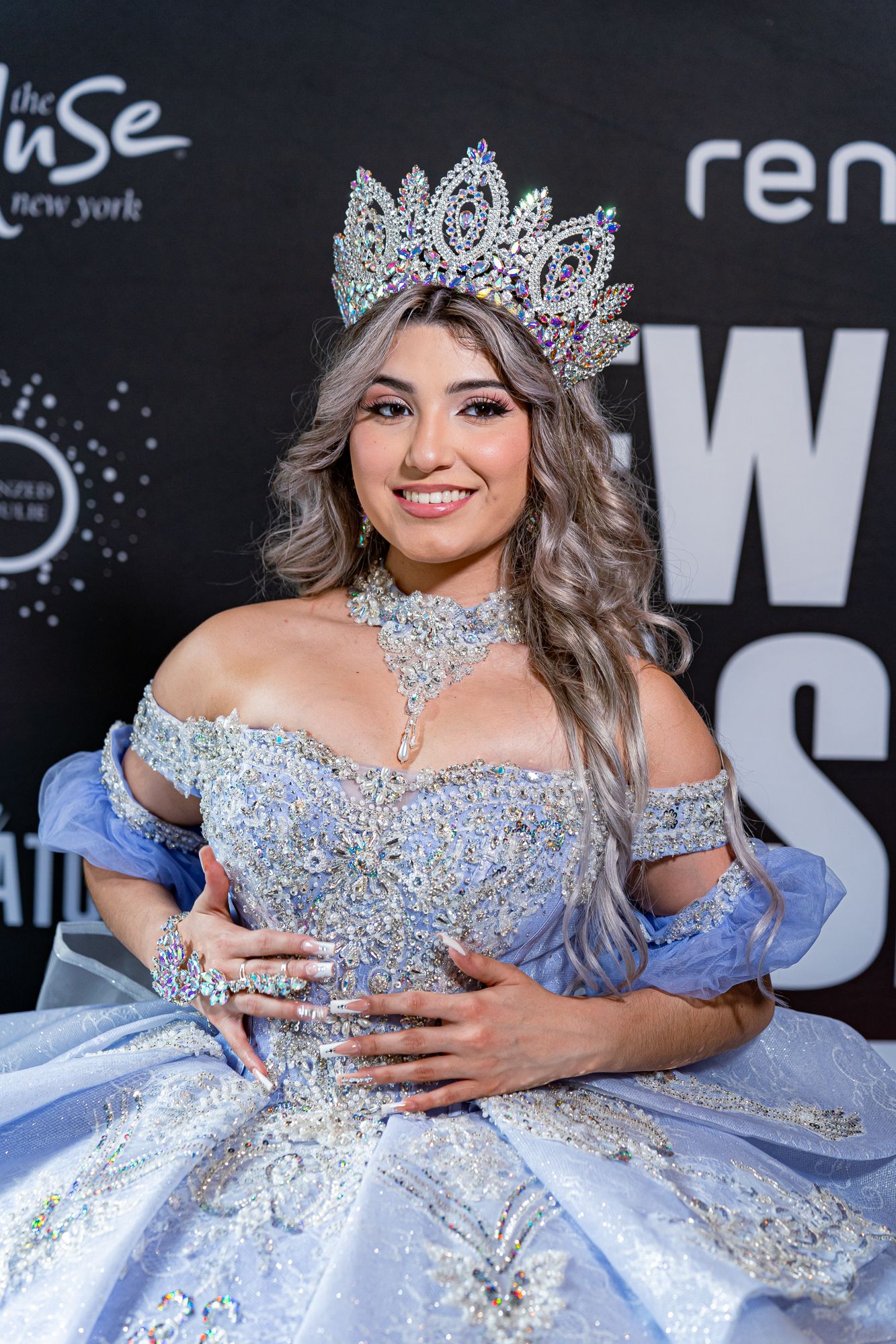 A beauty pageant contestant in a sparkling blue ball gown and crown poses on the red carpet at an event.