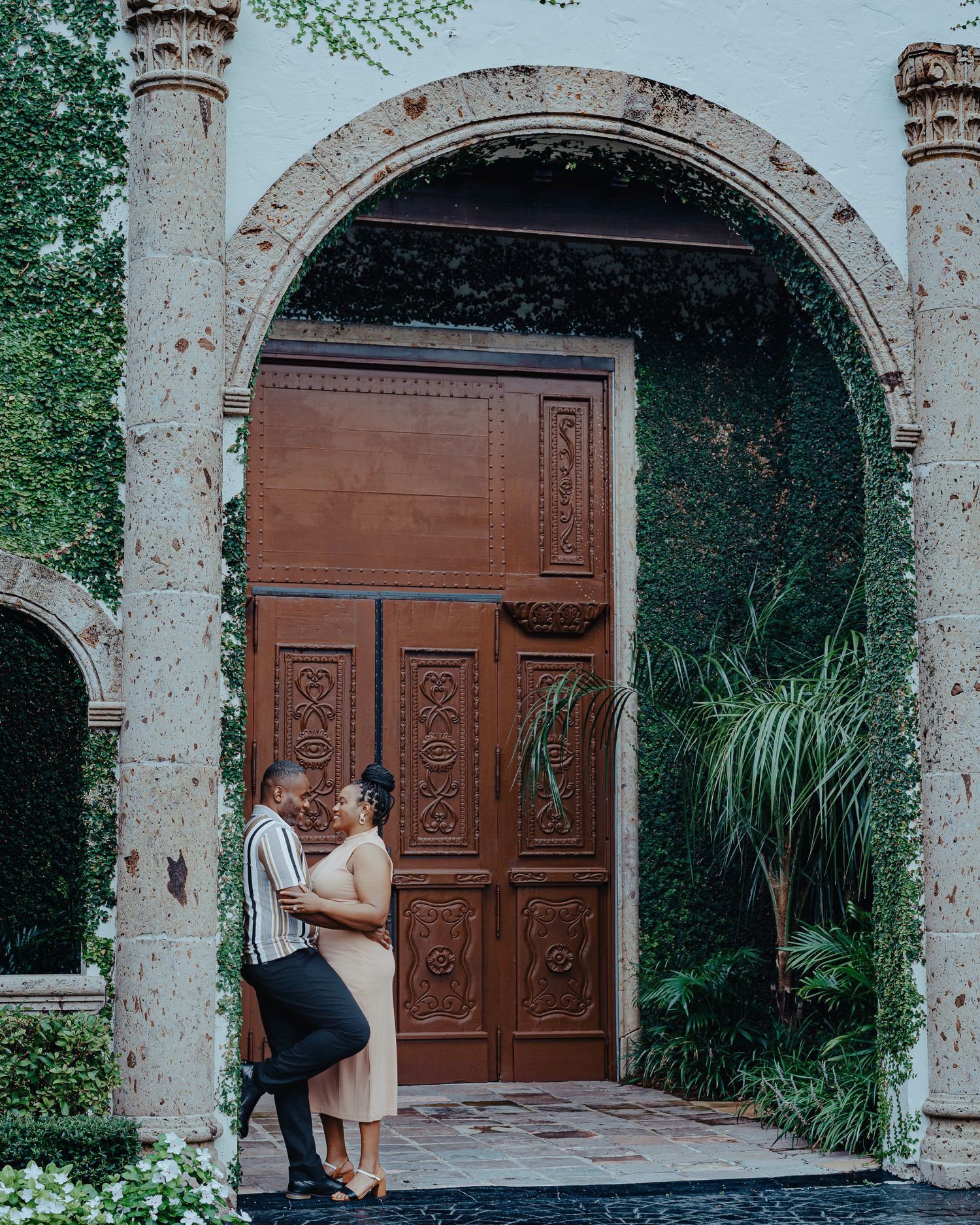 Couple embracing under ornate stone archway with wooden door and greenery.