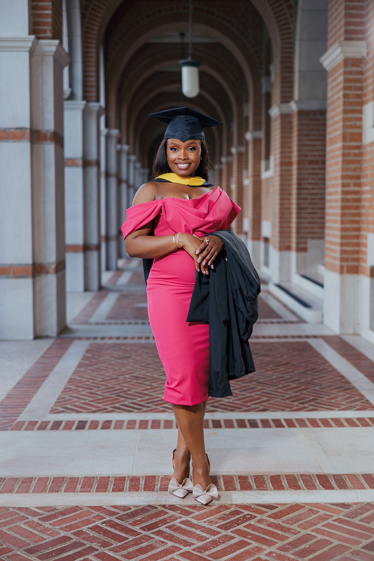 Graduate in pink dress and graduation cap poses in brick archway corridor for professional portraits.