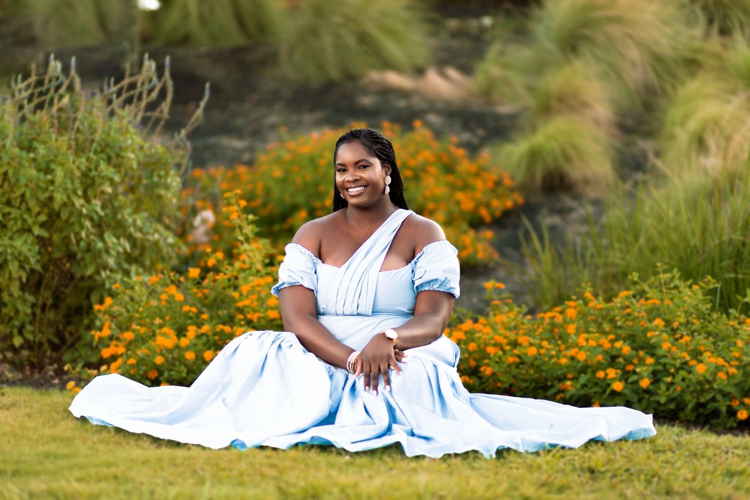 A person in a flowing white dress sits gracefully on grass surrounded by vibrant orange wildflowers and ornamental grasses.
