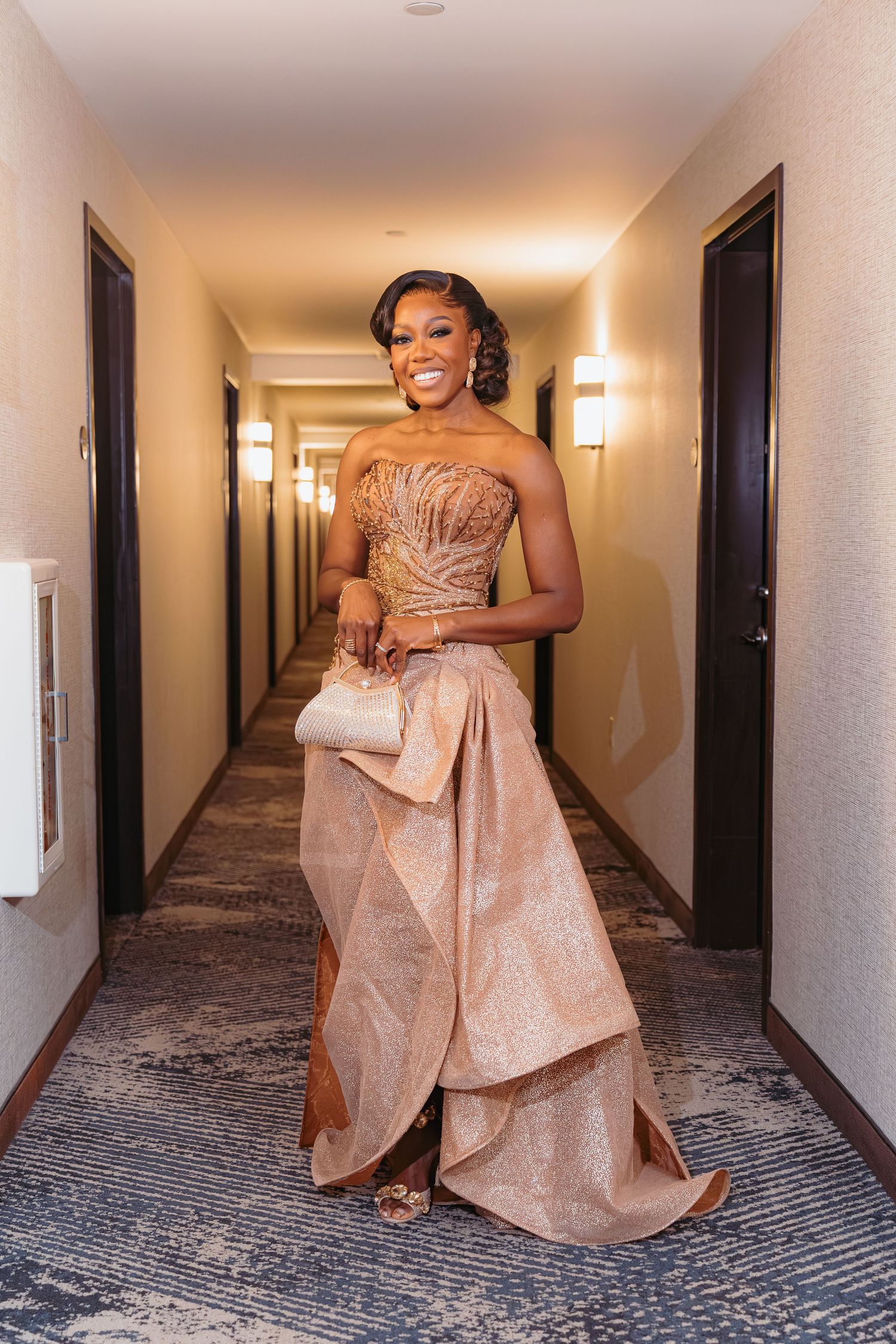 Someone poses elegantly in a rose gold strapless evening gown with dramatic ruffles in a hotel hallway.