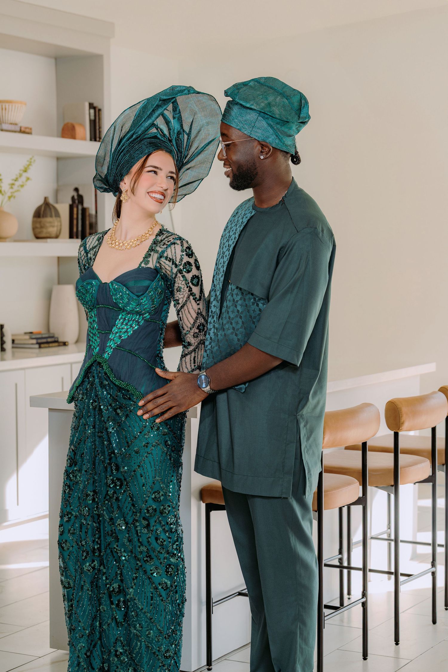 A couple wearing coordinated teal traditional outfits with matching headpieces share a joyful moment indoors.