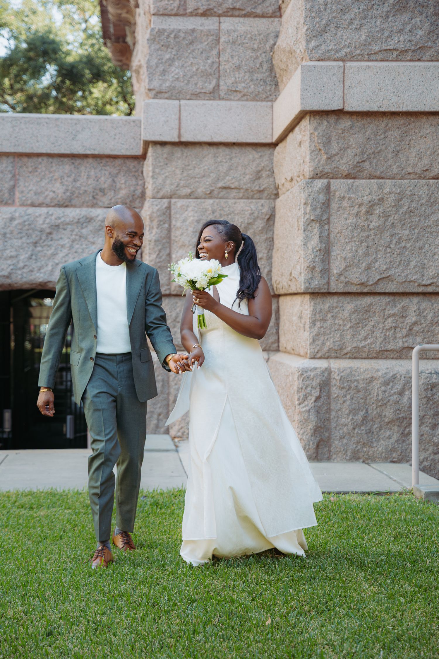 A couple in wedding attire walks together on grass near a stone building wall.