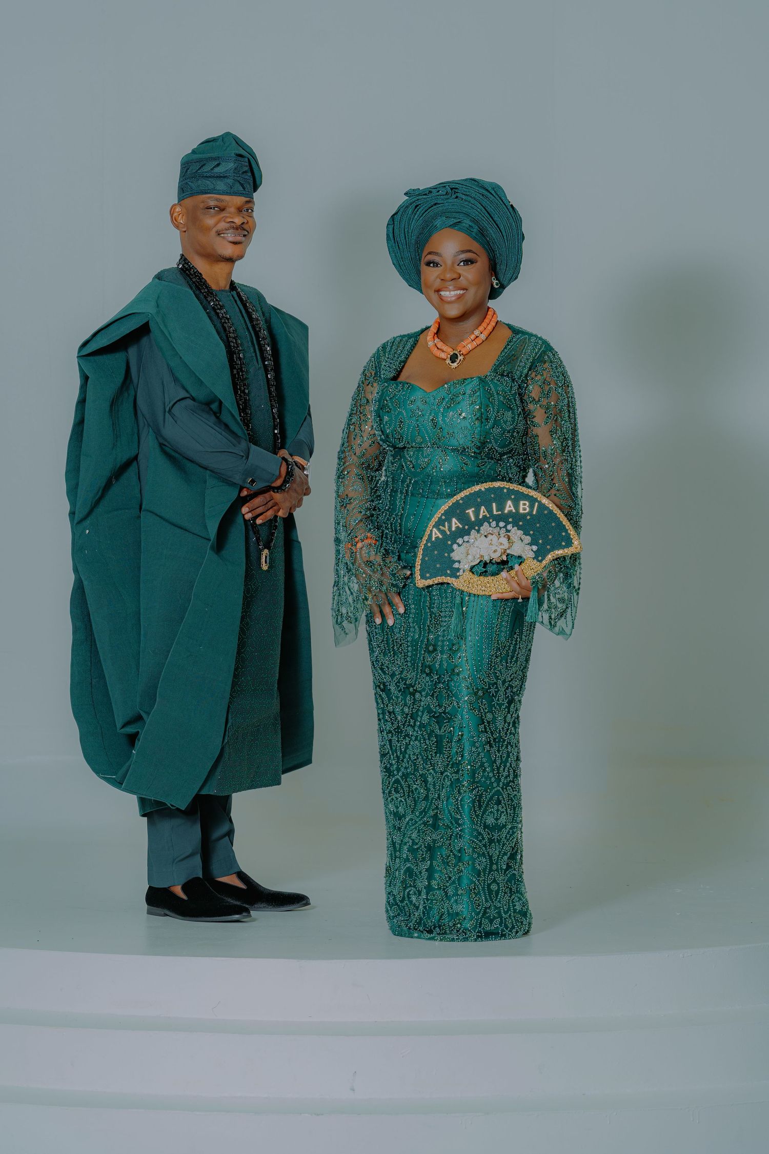 Nigerian couple in matching teal traditional attire, woman holding decorative fan, posing for formal portrait photo.