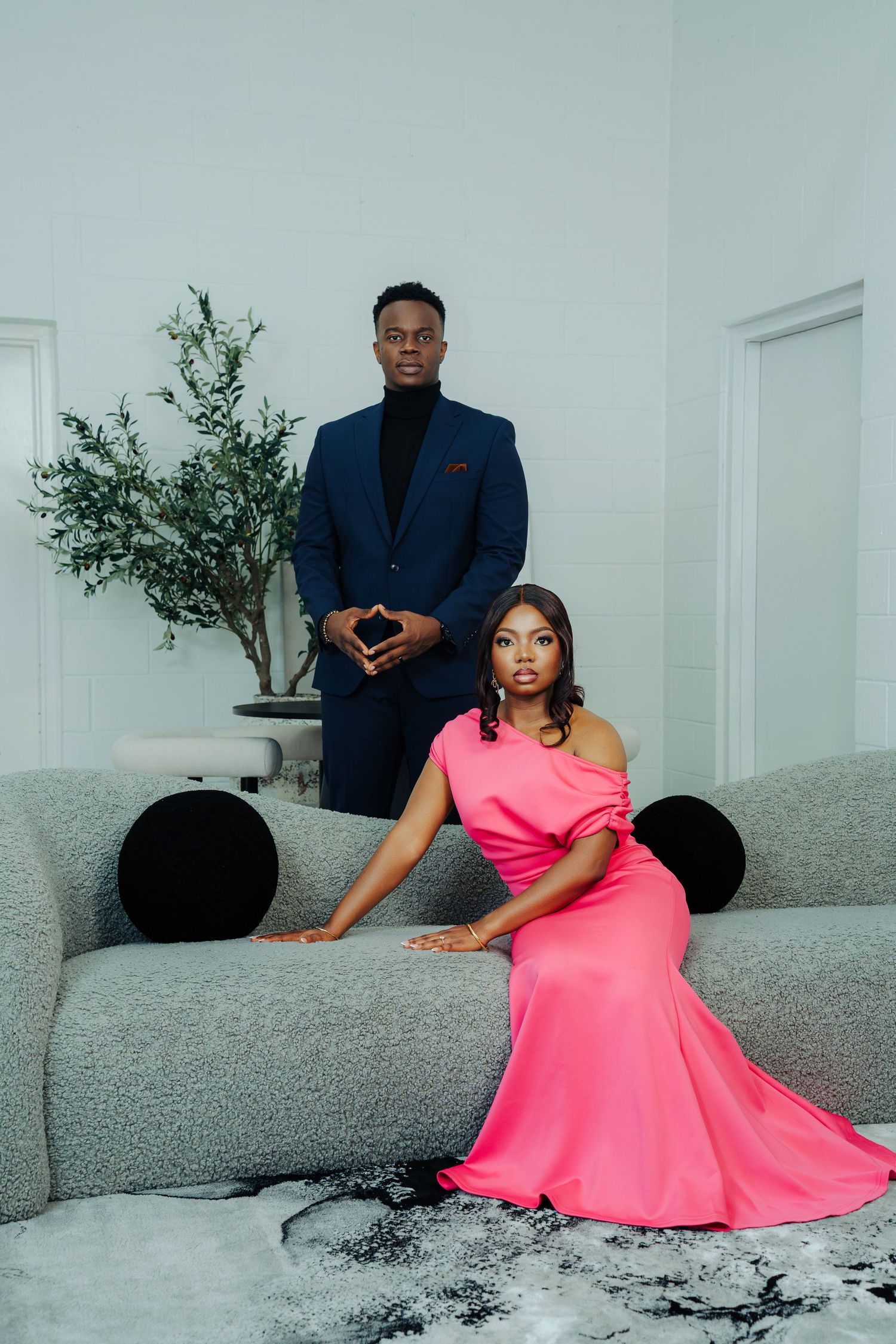 Elegant couple poses on a gray couch with a pink dress and navy suit for professional photos.
