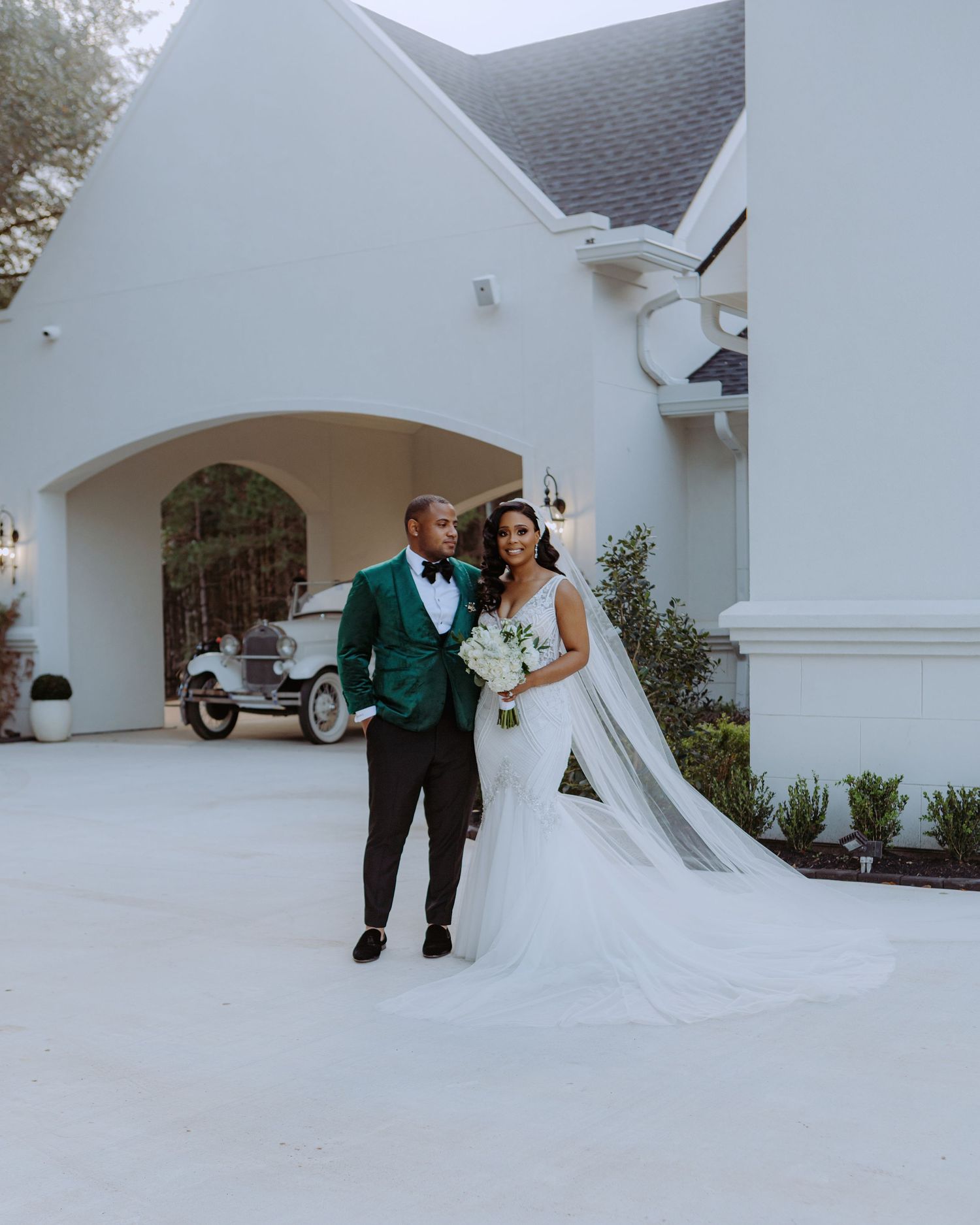 Wedding couple in emerald and white attire outside white chapel with vintage car.