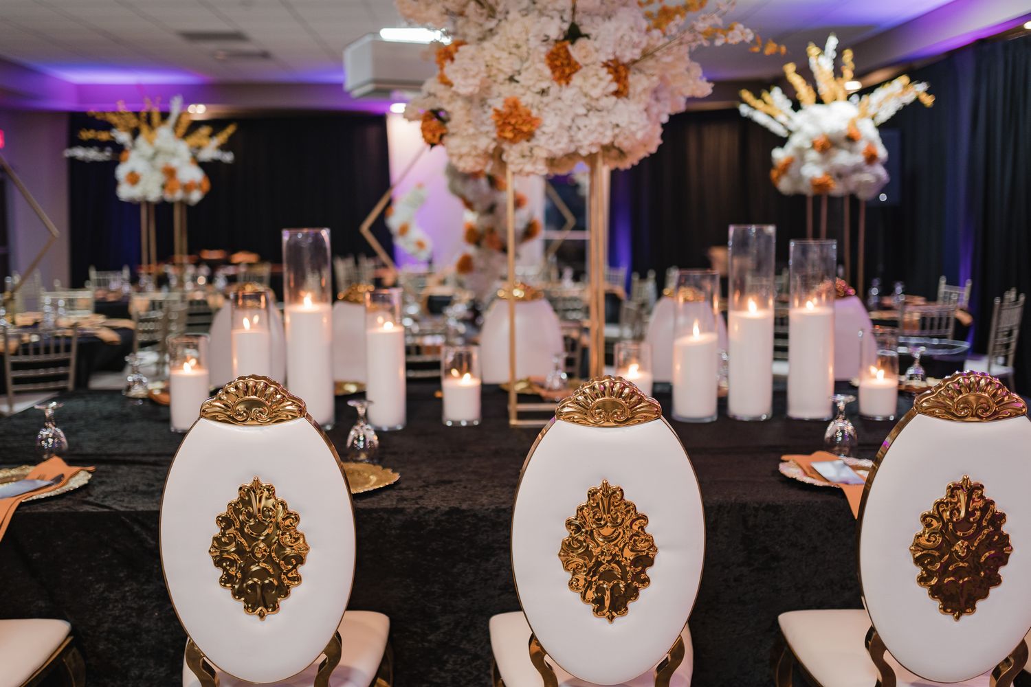 Elegant wedding reception decor features white centerpieces with gold accents and purple uplighting in a dark ballroom.