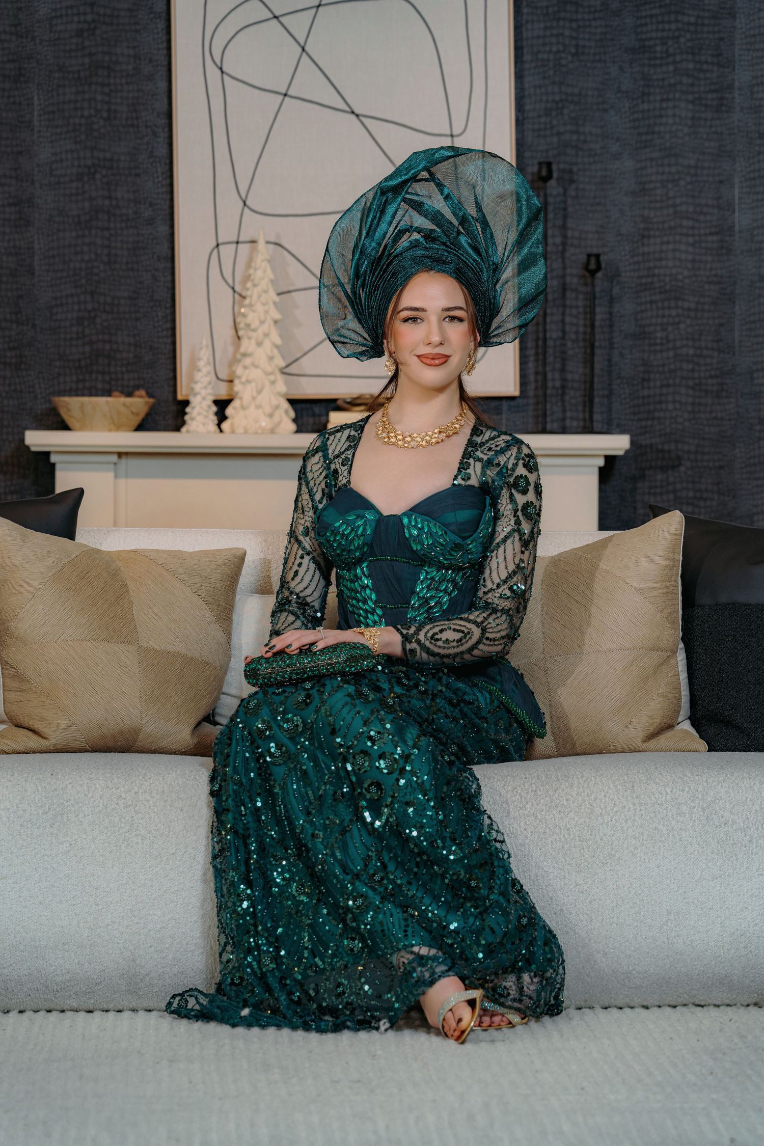 A person in an elegant emerald green sequined dress sits gracefully on a white couch against a dark wall.
