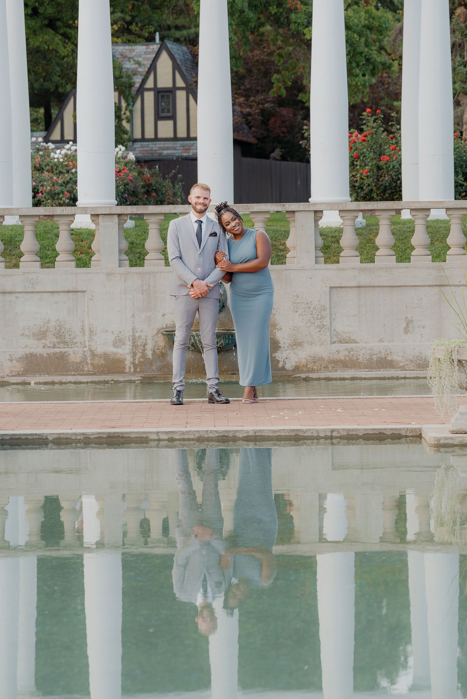 A couple poses together by a reflecting pool with white columns at a historic garden venue.